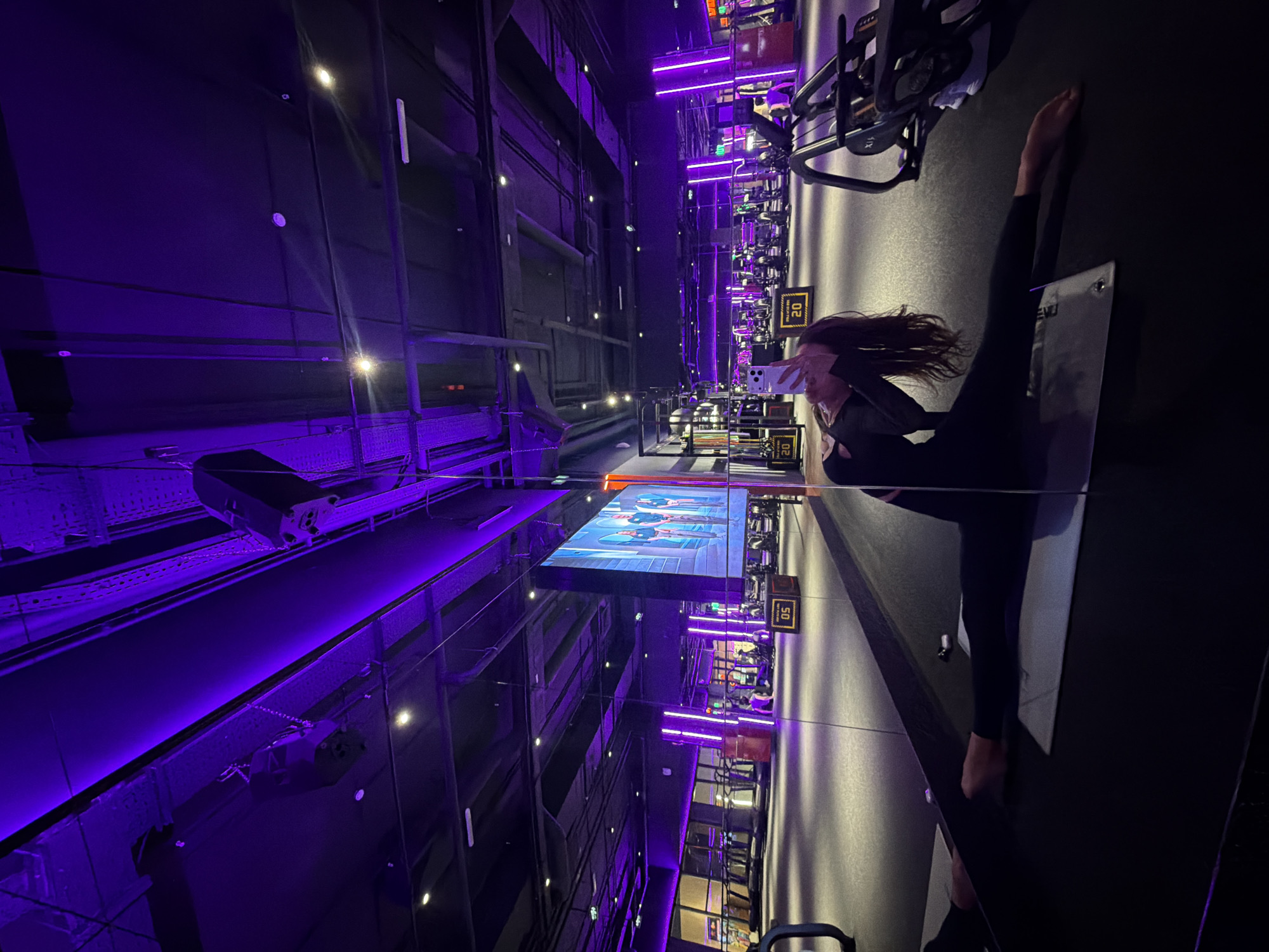 The image shows the interior of a gym. A woman in dark workout clothes stands on a mat, appearing to take a selfie with a phone. One leg is raised straight up. In the background are rows of stationary bikes and a large screen showing a workout class. Purple lighting illuminates the room. The scene is mirrored in the ceiling and walls, creating a vertical visual effect. Based on the limited information available in the prompt, the location in the city of Krasnoyarsk, Russia cannot be verified.