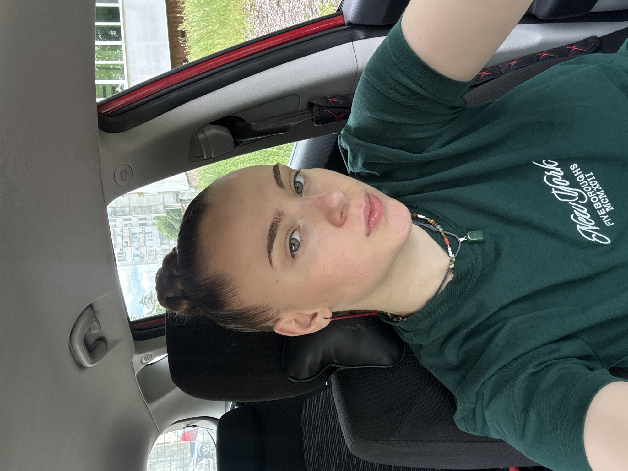 A young woman with her hair in a bun is sitting in the driver's seat of a car, taking a selfie. She is wearing a dark green t-shirt with a "New York Five Boroughs MCMXCII" graphic. She has a beaded necklace with a green pendant. The car's interior is visible, including the black headrest and seatbelt. Outside the car, a building and trees are visible. There is no indication of the specific location within Gabrovo, Bulgaria.