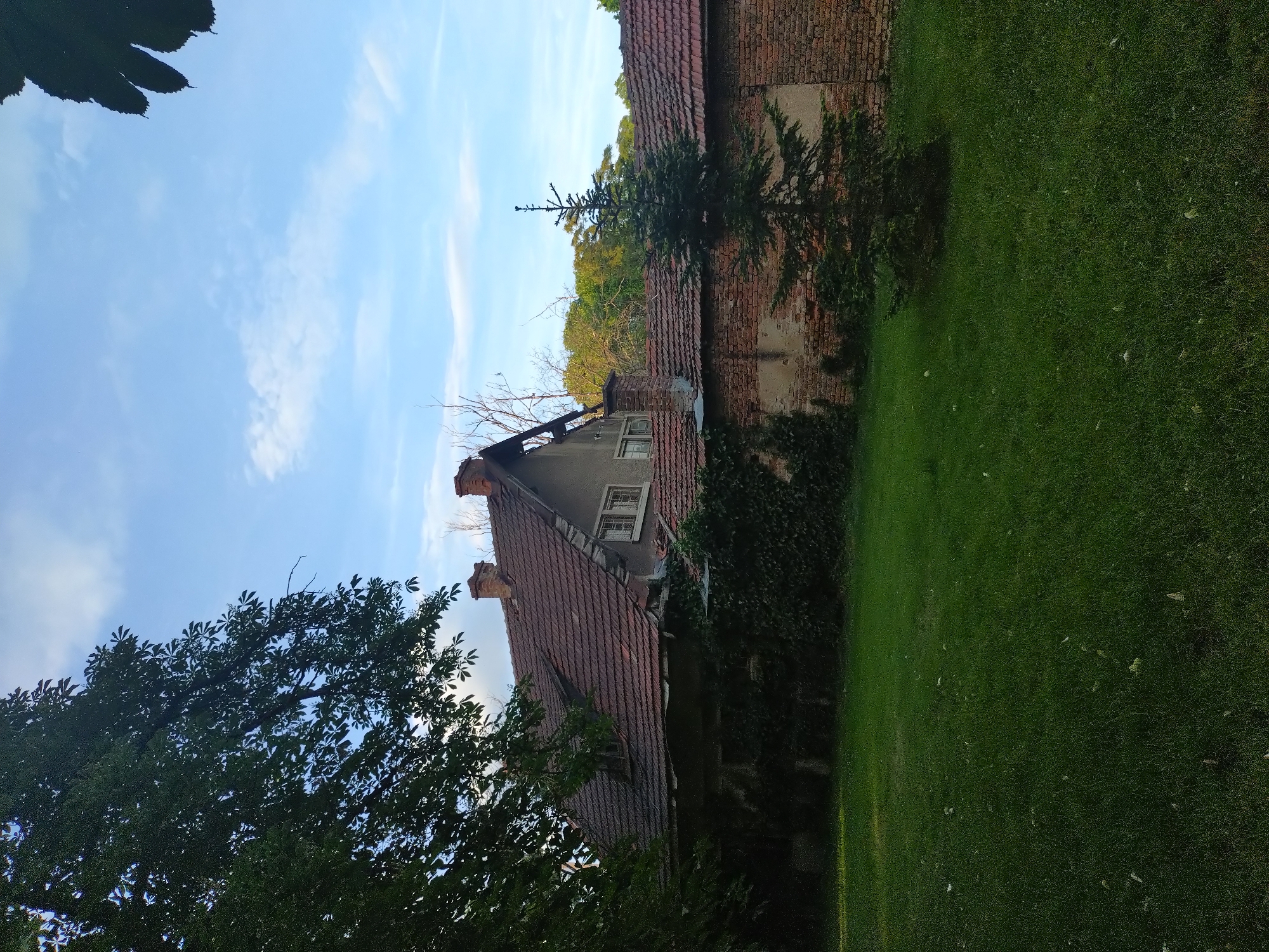 A medium shot shows a weathered building in Stara Zagora, Bulgaria, captured on a sunny day. The building features a red-tiled roof and is partially covered in ivy. Adjacent to it is a brick wall with a small tree growing near its base. A grassy lawn stretches out in the foreground, with scattered leaves. To the left, a large tree partially obscures the view of the building. The sky is blue with thin clouds.