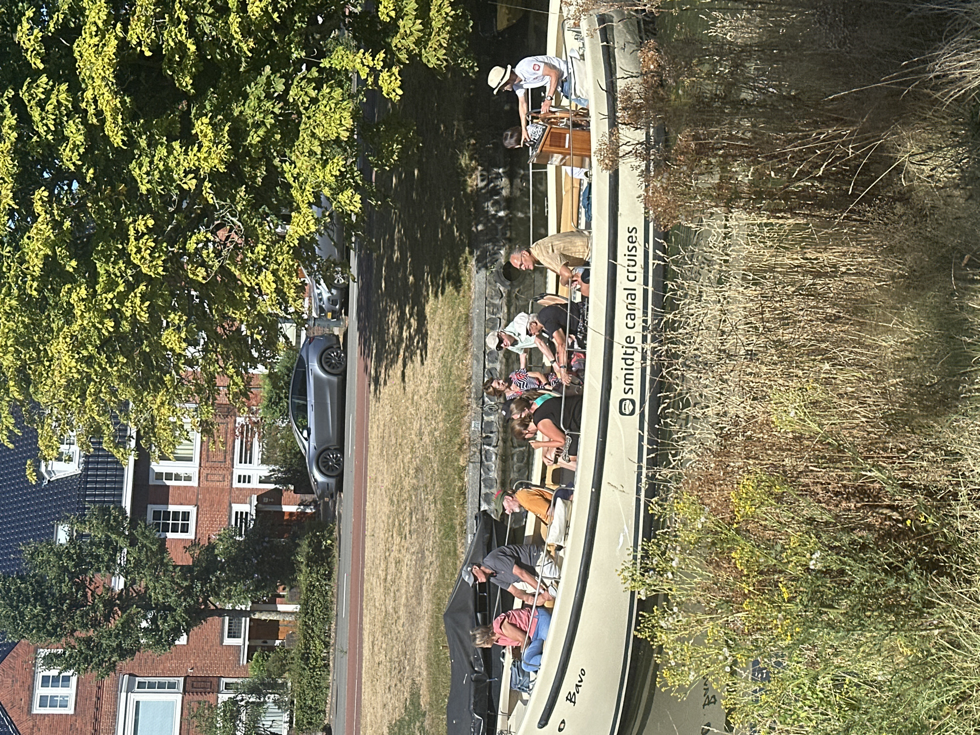 A boat with several passengers is navigating a canal in Haarlem, Netherlands. The boat is a light-colored vessel with a dark stripe along its side, and it has "Bavo" and "Smidje Canal Cruises" printed on it. The passengers appear to be enjoying a leisurely tour, with some looking at the scenery and others conversing. The boat is moving from left to right in the frame. The canal is flanked by a grassy embankment on the right, with lush green trees and vegetation. In the background, a residential street is visible, lined with brick houses and parked cars. A silver electric car is parked on the street, and another dark-colored car is partially visible behind it. The buildings have traditional Dutch architecture with dark tiled roofs and white-trimmed windows. The scene is illuminated by bright sunlight, suggesting it is a pleasant day. The overall atmosphere is serene and picturesque, characteristic of canal cities in the Netherlands.