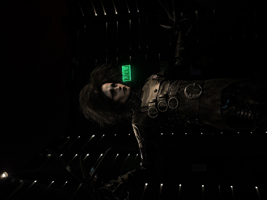 A dark, slightly angled shot features a person dressed as Edward Scissorhands. Their face is pale with dark hair, next to a glowing green exit sign. They wear a detailed, dark leather outfit with various straps and rings. Scissor-like hands are visible at the bottom and top of the frame. The background includes vertical light streaks. The overall scene is dimly lit, emphasizing the character and the eerie atmosphere.