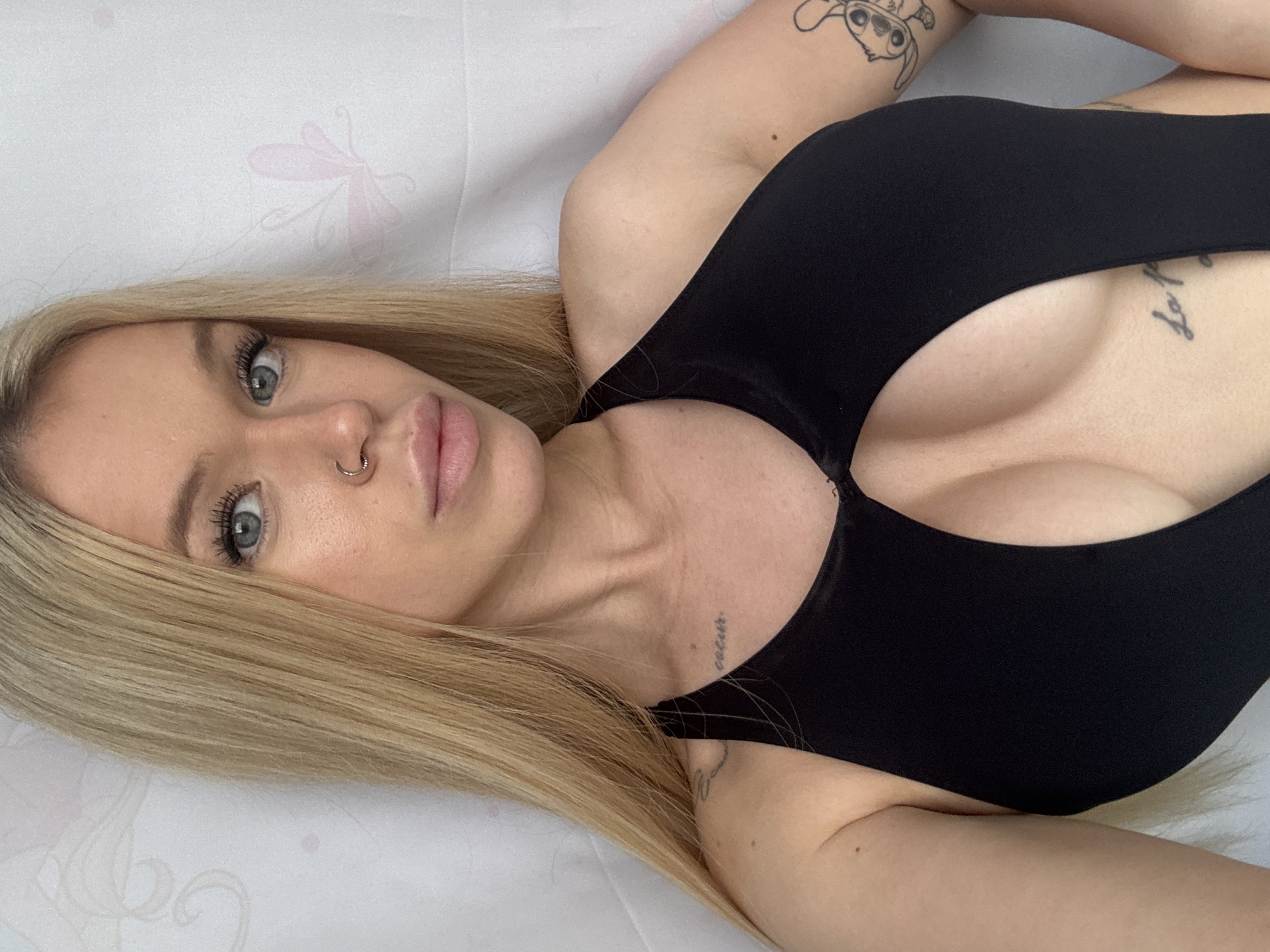 The image is a close-up, horizontal selfie of a young blonde woman lying down on a white surface with a subtle pink butterfly pattern. She is looking directly at the camera with a neutral expression. Her hair is long and straight, framing her face and falling across her chest. She has blue-grey eyes, full lips, and a nose ring. She is wearing a black top with a deep V-neckline that reveals her cleavage. There are visible tattoos on her left arm and shoulder, and a smaller tattoo on her collarbone. The lighting is soft, suggesting an indoor setting, possibly during the daytime. The overall mood is intimate and relaxed. There is no visible text in the image.