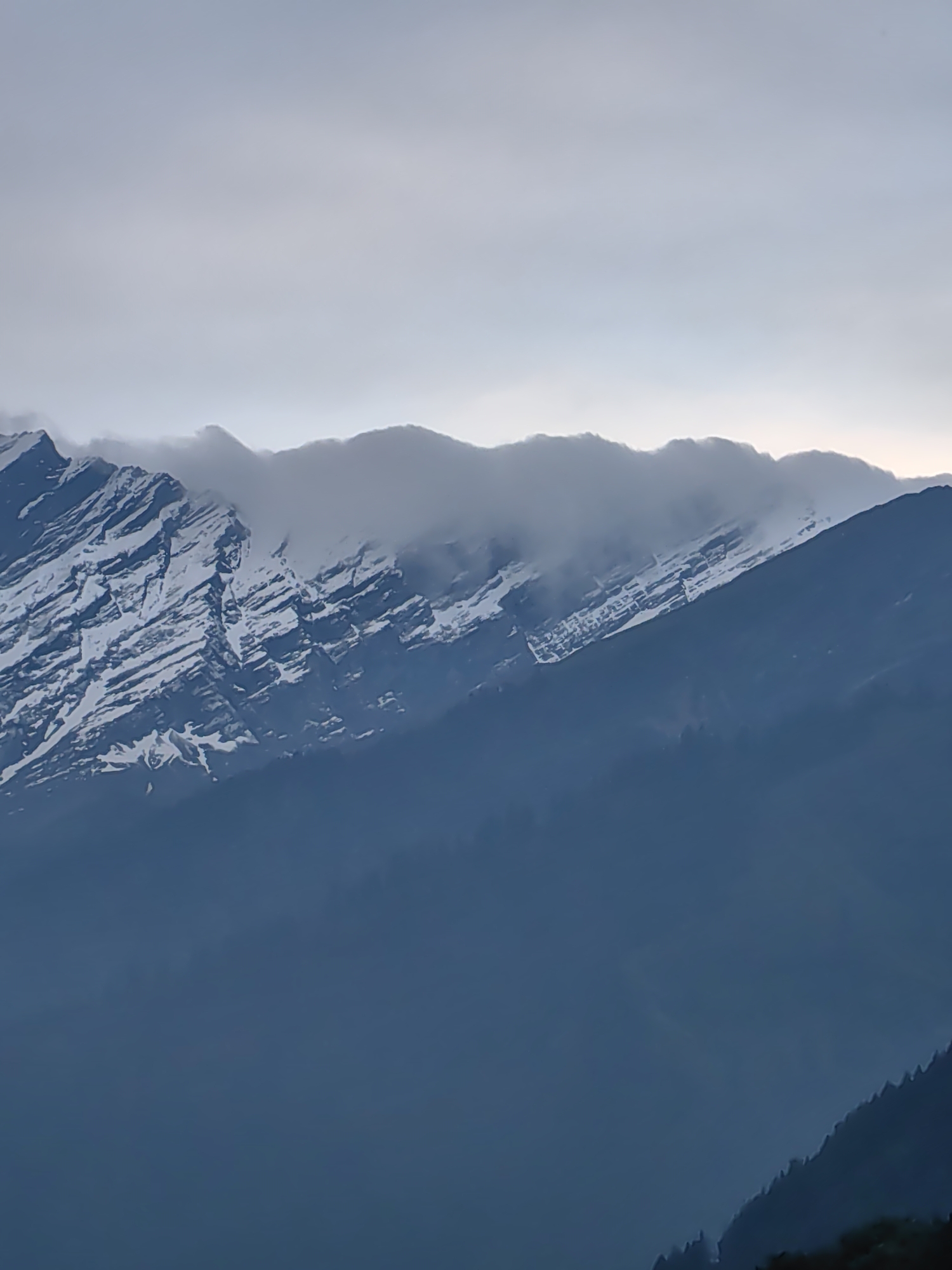 This image depicts a dramatic mountain landscape, likely taken in Vashisht, India. The scene is dominated by large, imposing mountains, with the peaks partially obscured by swirling clouds. The upper portions of the mountains are covered in snow, creating a stark contrast against the darker, shadowed slopes below. The texture of the snow-covered rocks suggests steep inclines and rugged terrain.
The overall atmosphere is serene and somewhat moody, hinting at the grandeur of nature. The time of day appears to be either early morning or late evening, given the diffused light and the soft, hazy quality of the sky, which transitions from a pale grey at the top to a darker, more muted tone closer to the horizon. The weather seems overcast, with the clouds adding a sense of mystery and depth to the scene.
In the foreground, the lower slopes of the mountains are covered in dark, dense foliage, possibly pine trees, which appear as a deep blue-black silhouette against the slightly lighter mountain range. The dense forest suggests a remote and natural setting. The composition of the image, with the layering of the distant snow-capped peaks, the mid-ground clouds, and the foreground forest, creates a sense of scale and vastness. There is no visible text or human activity in the image, emphasizing the raw and untouched beauty of the mountain environment.