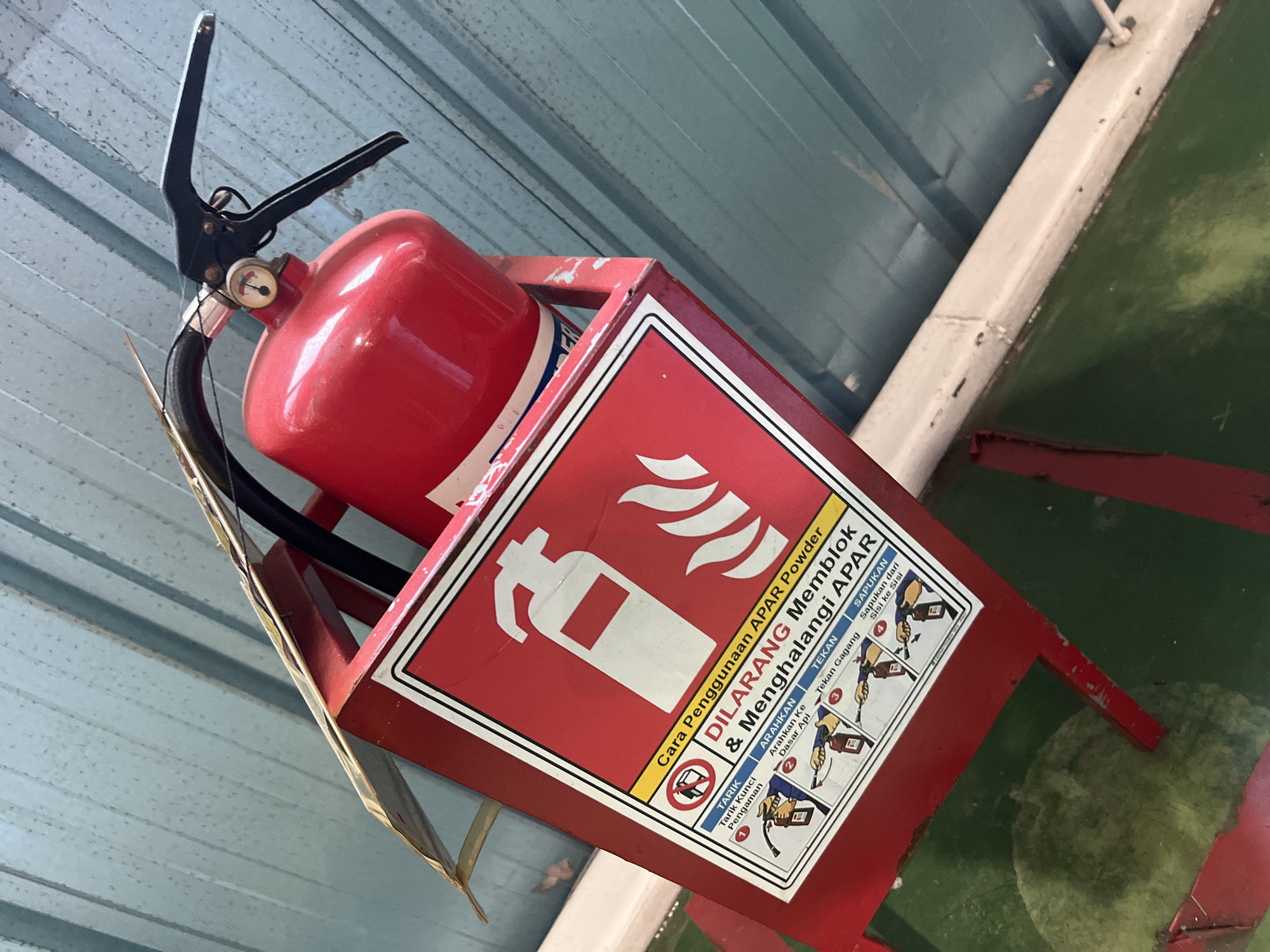 An overhead shot shows a red fire extinguisher mounted in a red metal bracket. The bracket is attached to a light blue corrugated metal ceiling. The fire extinguisher has a black handle and hose and a pressure gauge. A sign on the front of the bracket provides instructions for using the extinguisher in Malay, with diagrams illustrating the steps. The sign also states "DILARANG Memblok & Menghalangi APAR," which translates to "PROHIBITED to Block & Obstruct Fire Extinguisher." Below the bracket, a dark green surface is visible, possibly a wall or floor. The scene appears to be indoors.