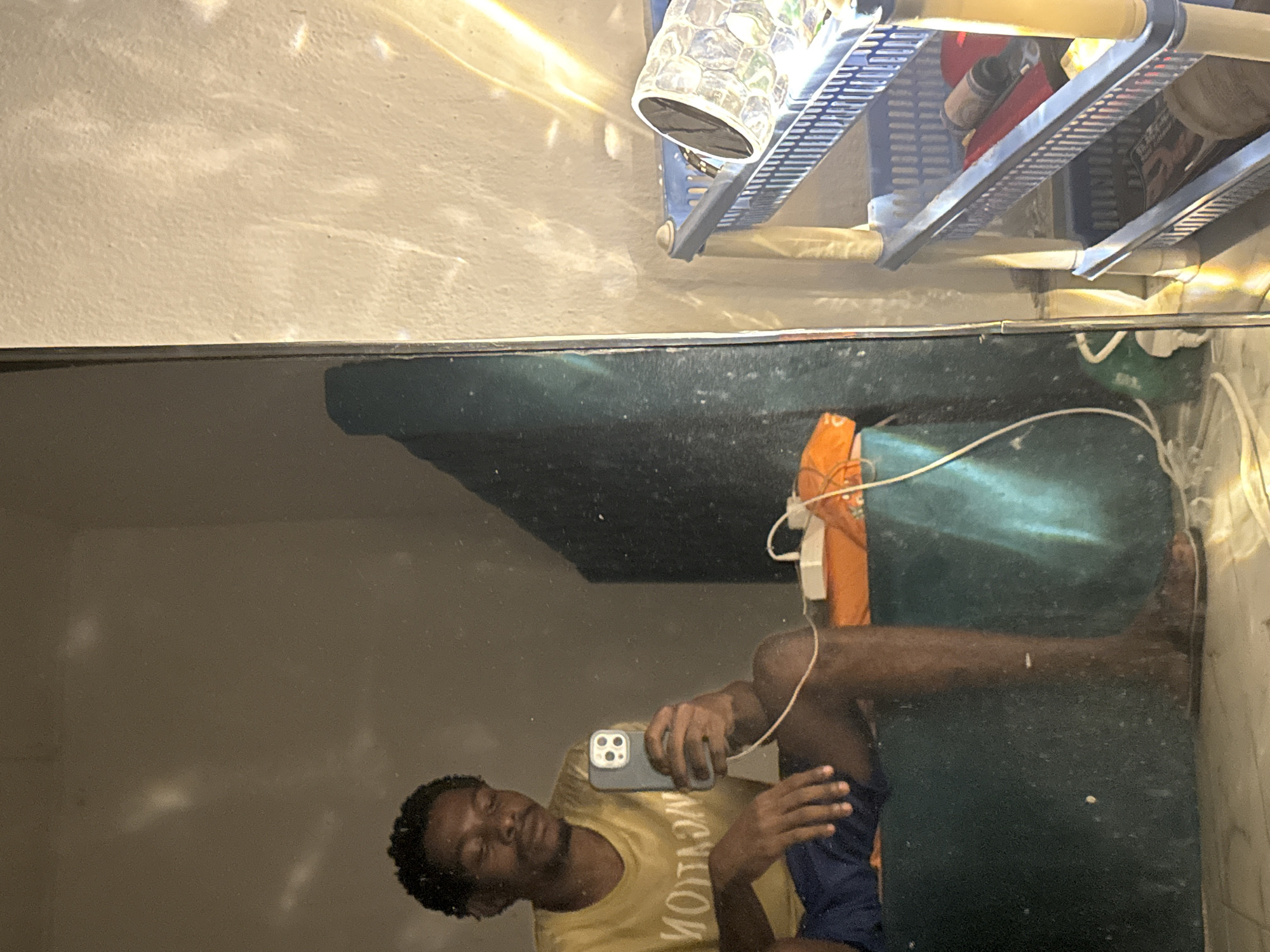 A young Black male is taking a mirror selfie in what appears to be a bedroom or living space in Uyo, Nigeria. He is seated, wearing a yellow t-shirt with the word "MOTION" visible in reverse, and dark blue shorts. He is holding a smartphone in his right hand and looking at it. To his right, there is a multi-tiered blue plastic shelf unit containing various items, including a clear decorative object, what looks like a flashlight or speaker, and some magazines or books. The background wall has a textured appearance and is illuminated by a strong light source casting patterns. Wires and an orange object are visible on the floor near the person. The overall environment suggests a casual, indoor setting.