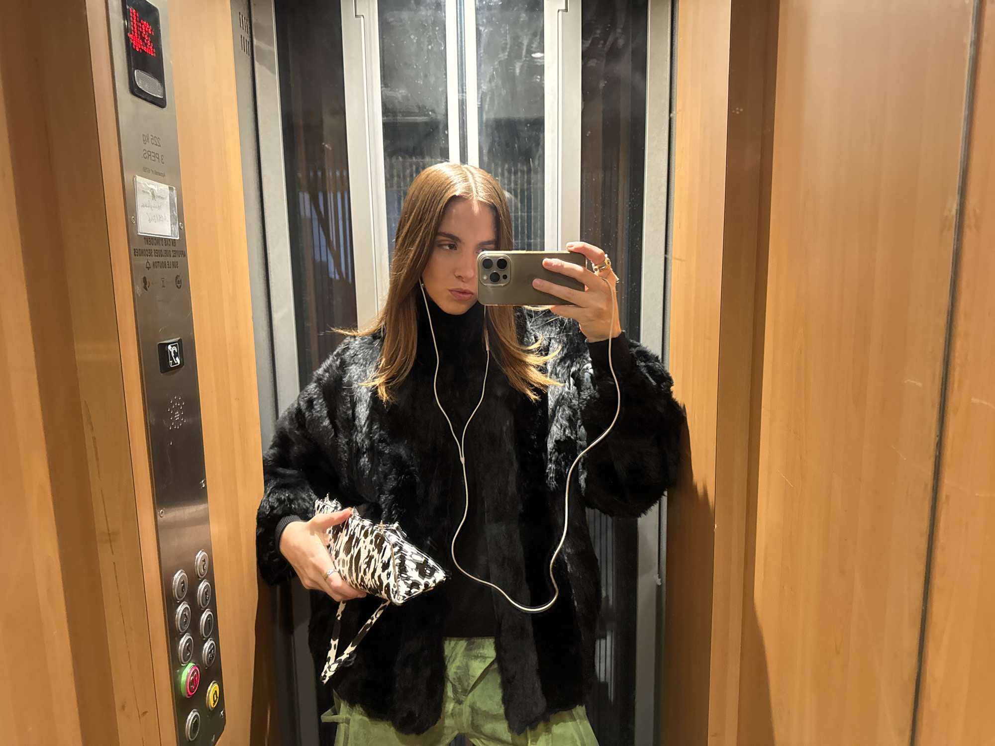 A woman takes a mirror selfie inside an elevator. The elevator's interior is paneled in light brown wood with a stainless steel control panel. She wears a black fur coat, a black turtleneck, and green pants. She holds a black and white patterned purse. The elevator doors are closed, partially obscuring a blurred view of the outside. The current floor is displayed as "13" on the control panel. Although the exact location cannot be determined from the image, elevators are common in commercial and residential buildings globally, including Paris, France.
