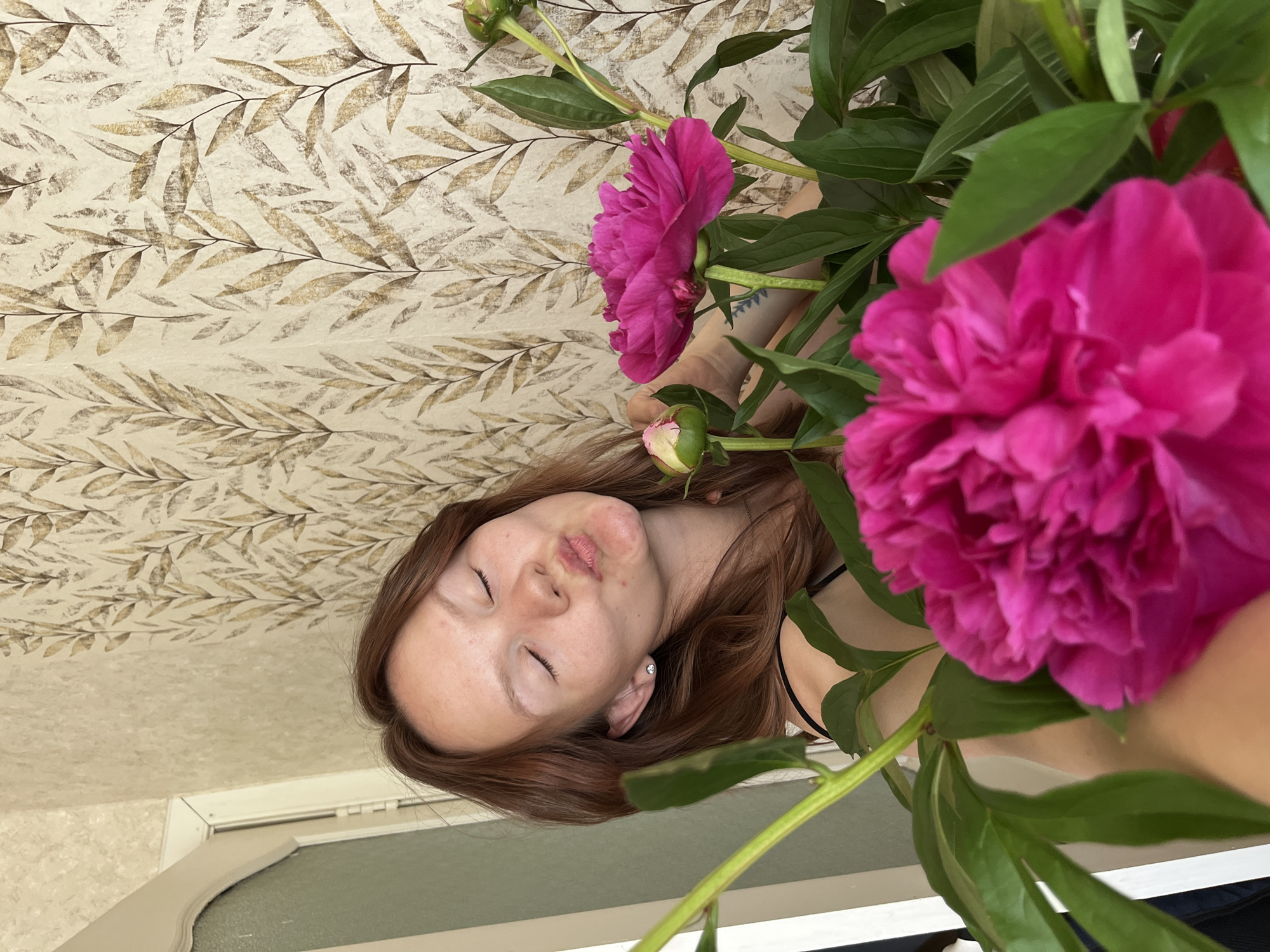 The image depicts a young woman with auburn hair, her eyes closed and lips puckered in a kiss, as she poses with a bouquet of vibrant pink peonies. She is wearing a black tank top and a small stud earring. The background features wallpaper with a delicate, repeating pattern of stylized leaves in shades of beige and brown, and a portion of a doorway to the left. The setting appears to be an indoor residential space. The photograph does not contain any discernible elements that would allow for its specific location within the city of Chaikovsky, Russia, or any other city.