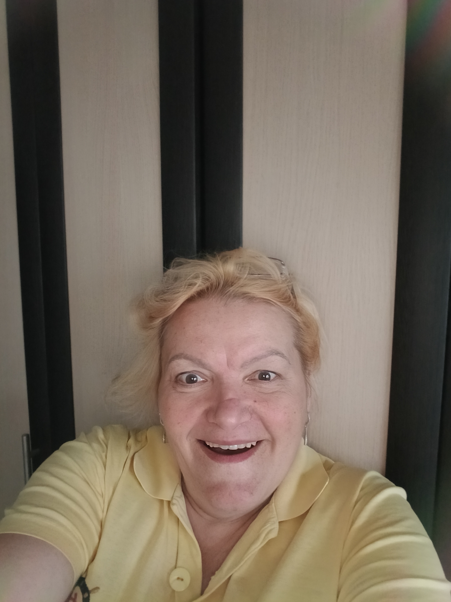 The image depicts a smiling middle-aged woman with short, blond hair, wearing a yellow collared shirt. She appears to be taking a selfie indoors. The background consists of a light-colored door with dark, vertical inlays. The photo's perspective suggests the woman is holding the camera slightly above her. The location is identified as Красный Маяк, Russia, but there is no visual information in the image to confirm the city.