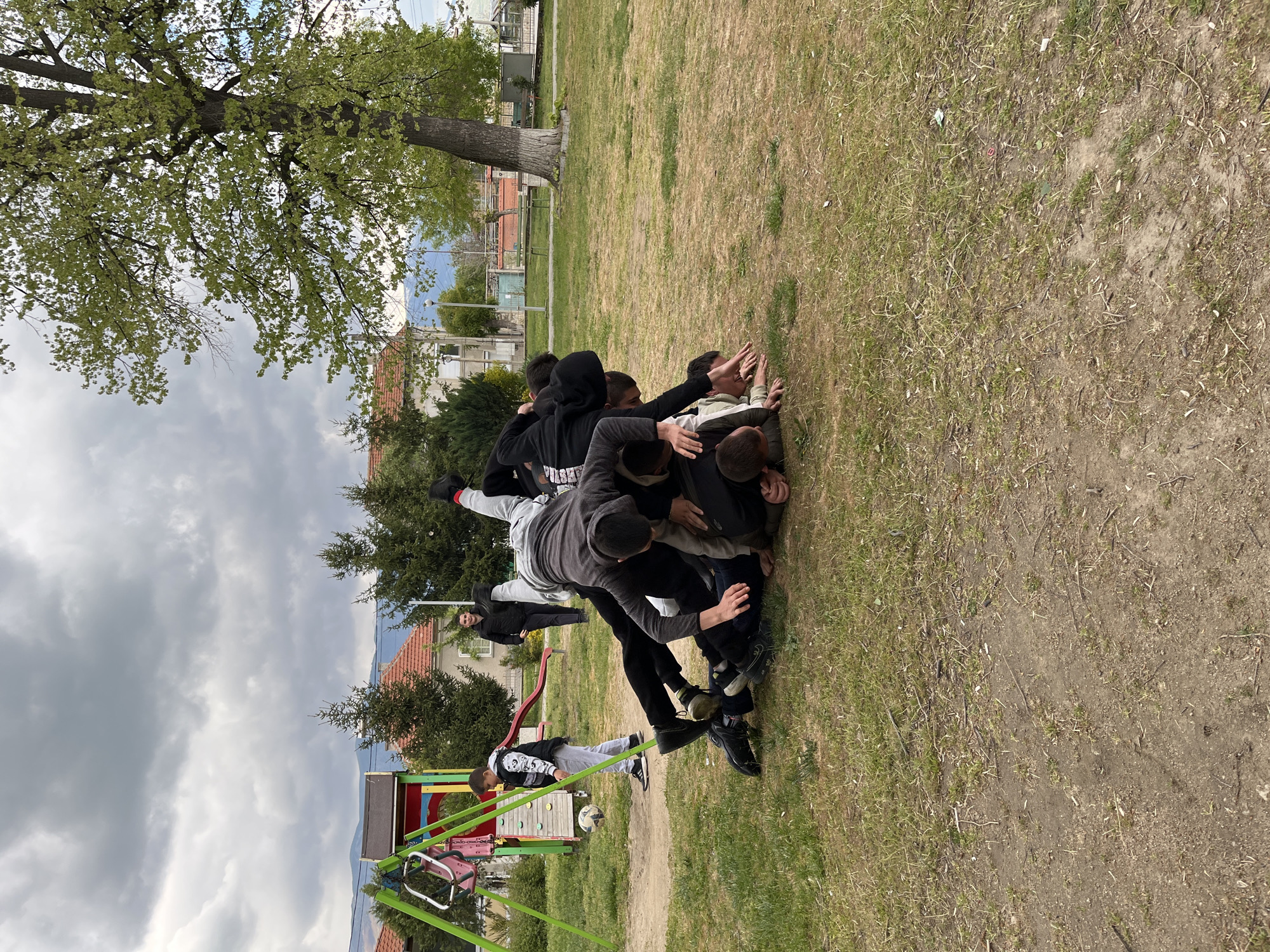 A group of approximately 10 young people are engaged in a physical altercation on a grassy area in Karavelovo, Bulgaria. Some are standing, while others are piled on top of each other on the ground. A playground swing set is visible in the foreground. A soccer ball is near the swing set. Buildings and trees surround the grassy area. The sky is overcast.