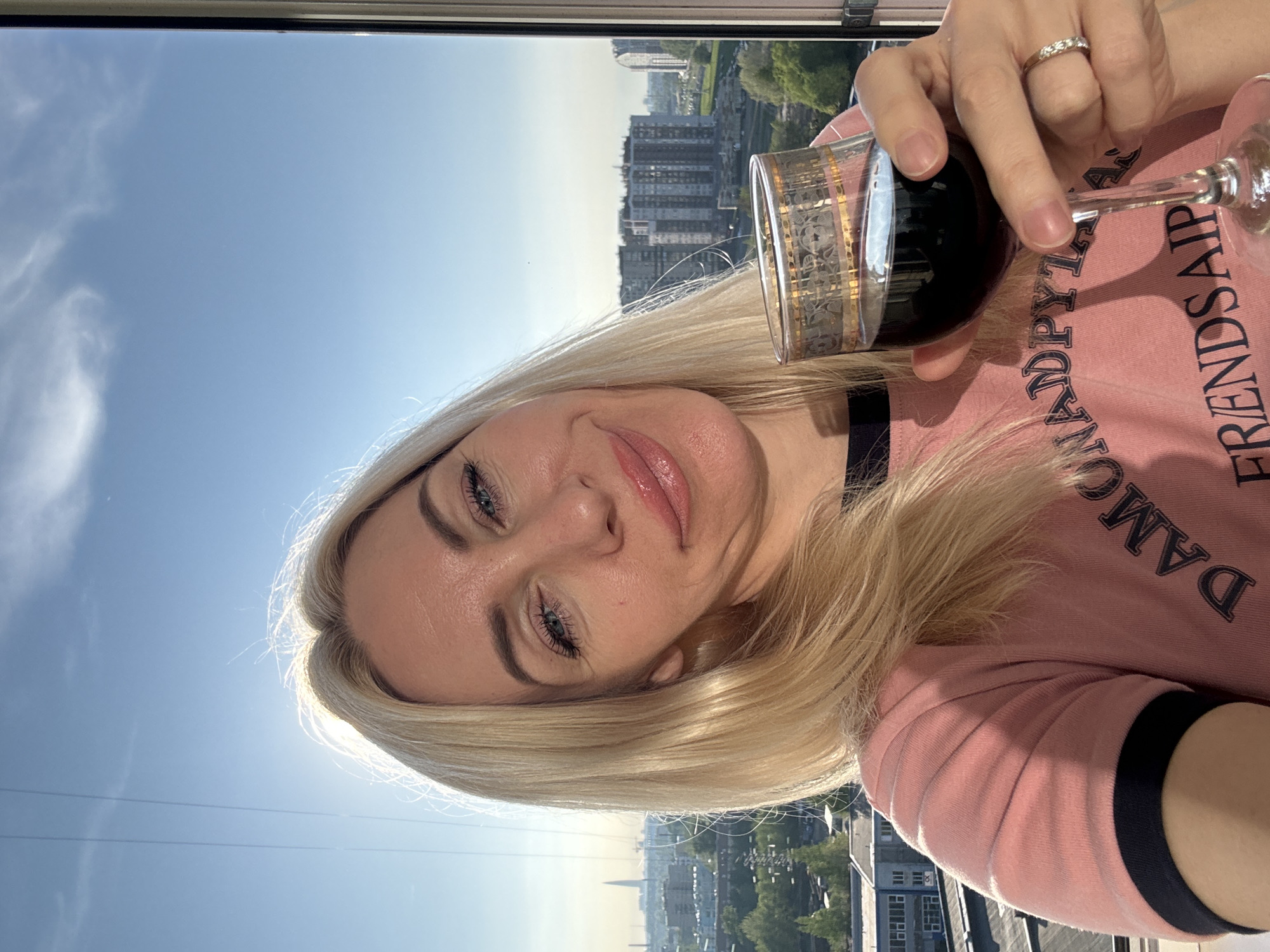 A fair-skinned woman with blonde hair holds a glass of dark liquid in front of a city scene. The city has tall buildings, trees, and a spire in the distance. The sky is blue with some clouds. The woman is wearing a pink long-sleeved shirt with black trim. She has a ring on her finger. The location of the city is Unknown, Unknown.
