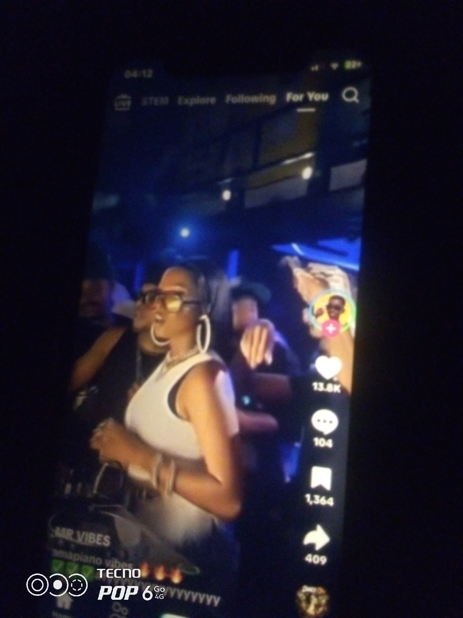 The image is a low-quality, vertically oriented photograph, likely taken with a mobile phone, capturing a screen displaying a social media application. The screen shows a video with several UI elements characteristic of platforms like TikTok or Reels.
The main subject is a woman in the foreground, wearing large hoop earrings, sunglasses, and a white tank top. She is positioned slightly off-center to the left. Her hands are visible, one appearing to hold something near her chest. In the background, other individuals are present in what appears to be a dimly lit indoor environment, possibly a club or event, with blue lighting visible.
On the right side of the screen, there are engagement icons and counts typical of social media platforms: a profile picture with a "+" symbol, a heart icon with "13.8K" likes, a speech bubble icon with "104" comments, a bookmark icon with "1,364" saves, and a share icon with "409" shares.
At the top of the screen, a time display reads "04:12" and navigation options are visible including "STEM," "Explore," "Following," and "For You." There is also a search icon.
In the bottom left corner of the screen, text overlays read "MR VIBES," "amapiano vibes," and "TECNO POP 6 GO 4G yyyyyyyyyy." This suggests the video is related to music and potentially promoting a mobile device.
The overall scene is dark with the illuminated screen being the primary light source. The image quality is poor due to low light conditions and potential motion blur, making fine details difficult to discern.
There is no visual information in the image that directly indicates the location as Howayi, Nigeria. This detail appears to be external information provided in the prompt.