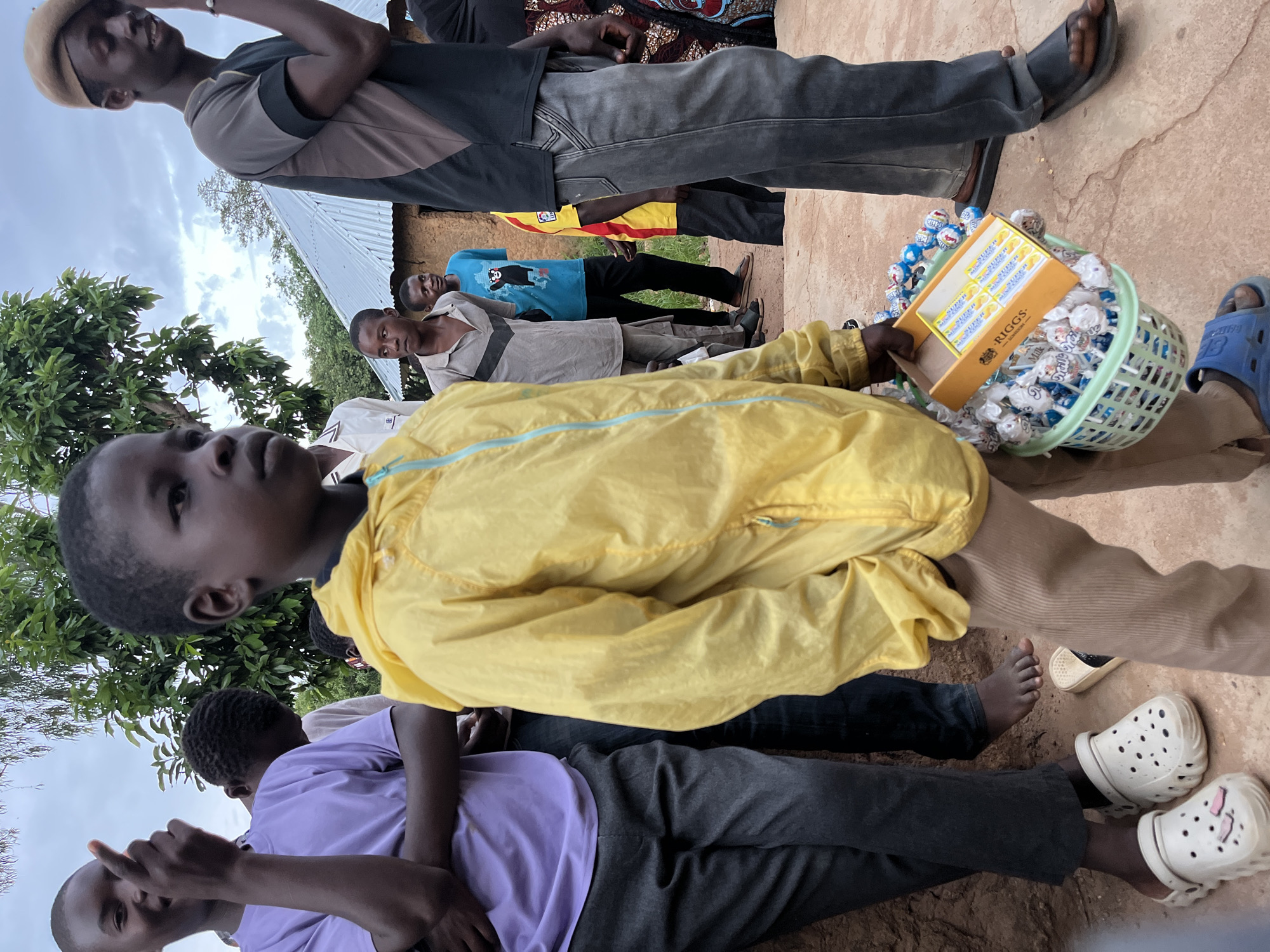 The image depicts a group of young people gathered outdoors, possibly in a village setting near Jos, Nigeria. In the foreground, a young boy wearing a bright yellow jacket is holding a green plastic basket filled with lollipops. A box of "RIGGS" candy is visible on top of the lollipops. The boy is looking away from the camera. Behind him, other individuals are visible, including a person in a light purple shirt and dark pants, and another in a grey shirt. In the background, a building with a corrugated metal roof and earth walls can be seen, suggesting a rural or semi-rural environment. The sky is overcast.