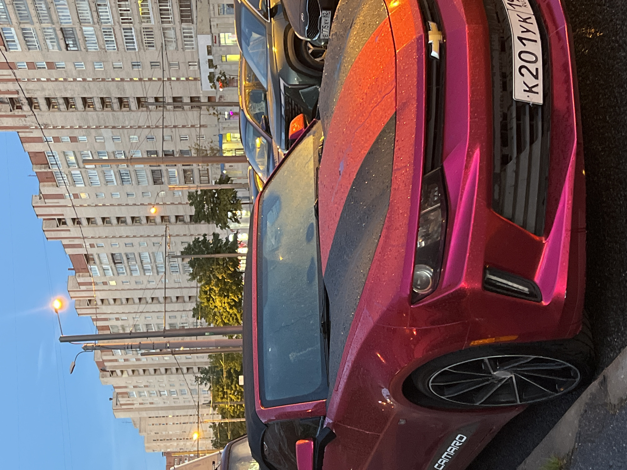 This media file captures a close-up, angled view of a red and black Chevrolet Camaro parked on a street in Saint Petersburg, Russia. The car is the primary subject, with its distinctive paint job, including black racing stripes on the hood and roof, and a metallic pinkish-red hue. The vehicle appears to be wet, suggesting recent rain or a wash. The front of the car, including the grille, headlights, and a portion of the wheel, is clearly visible. A license plate with Cyrillic text is affixed to the front.
In the background, a multi-story residential building with numerous windows stretches across the frame. The building's facade is detailed with brickwork and balconies, typical of Soviet-era architecture. Streetlights and power lines are also present, indicating an urban environment. Trees with yellow-green foliage are visible between the building and the car, suggesting it is either spring or autumn. The sky is a clear, pale blue, hinting at either early morning or late afternoon.
The scene depicts a static moment, likely during a period of rest or parking. There are no people visible in the frame. The lighting and atmosphere suggest an overcast day or twilight. The visible text on the license plate reads "K201YK" and "RUS", confirming its Russian origin. Another piece of text, "CAMARO", is visible on the lower part of the car. The overall impression is one of urban street photography, focusing on the details of the car against the backdrop of a residential cityscape.
