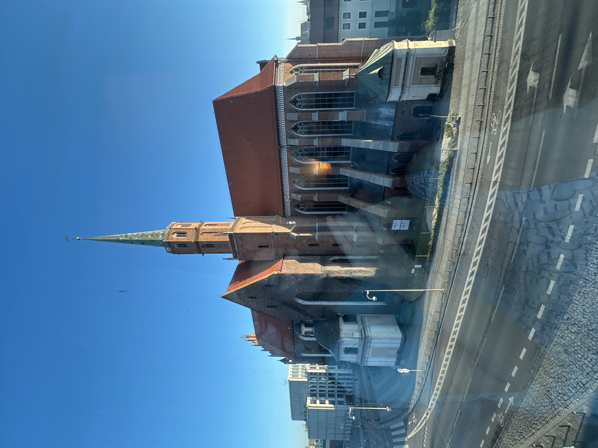 Here is a description of the image:
The image shows a red brick church with a tall, green-topped spire in Wrocław, Poland, seen from an elevated viewpoint, possibly a window. The sky is a clear blue. The church is adjacent to a street with clearly marked bike lanes and pedestrian crossings. Nearby are modern buildings, some appearing residential and some commercial. The scene is bright, suggesting a sunny day. Reflections are visible on the glass, indicating that the photograph was taken from indoors. The perspective is angled, giving a partial view of the rooftops of buildings across the street. The architecture of the church is Gothic, with prominent arches and detailing.