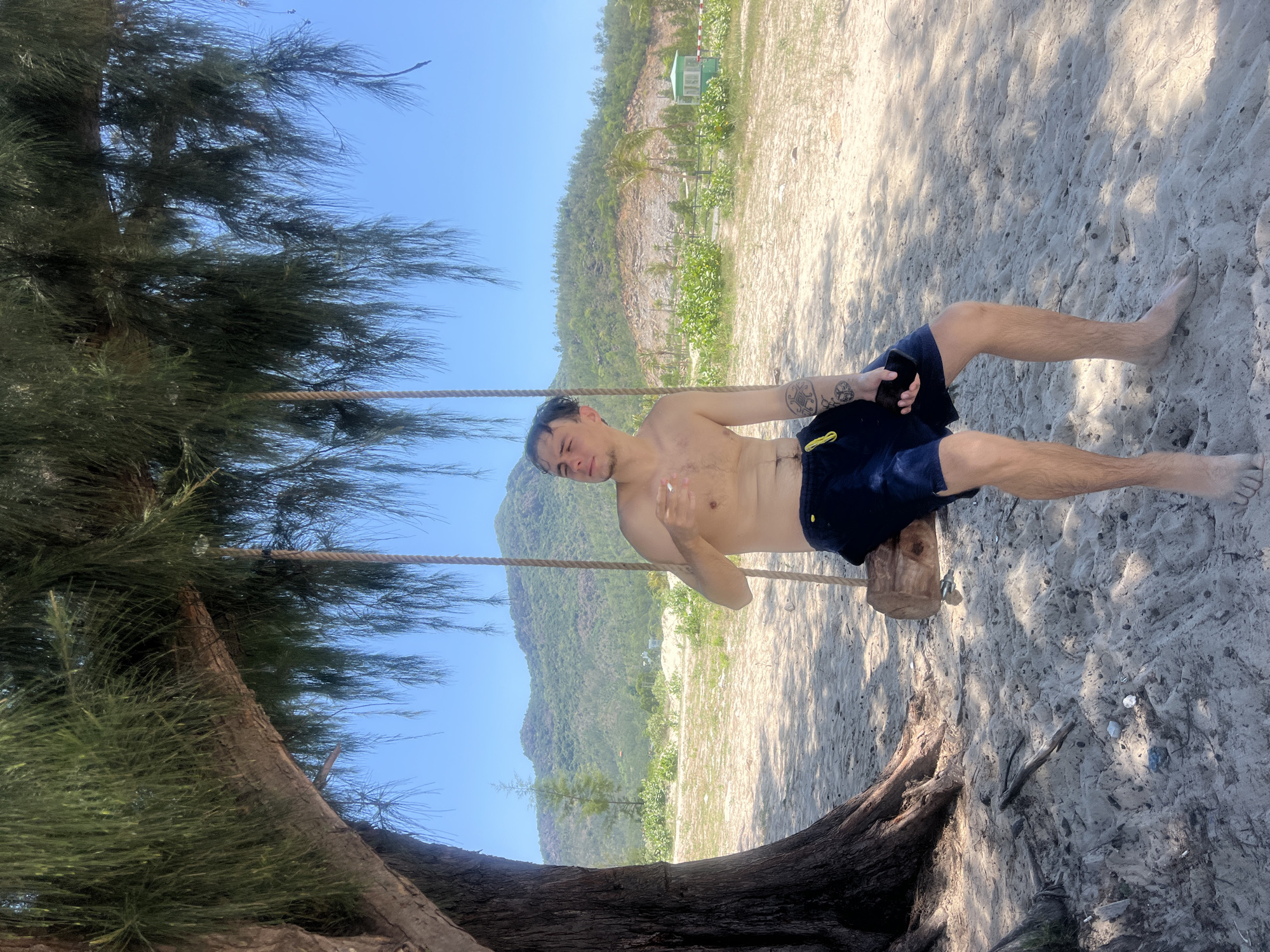 A young Caucasian man is sitting on a wooden swing suspended from a tree branch, on a sandy beach. He is shirtless and wearing dark blue shorts. His left hand is holding a cigarette and his right hand is holding a dark colored smartphone. He is looking to his left, with his eyes closed and a slight smile on his face. The swing is made of a thick piece of wood and is suspended by two ropes. The tree is large and has long, thin needles. The background shows a hilly landscape with sparse vegetation and a bright blue sky. There is a small green structure visible in the distance, possibly a lifeguard station or a small building. The beach is made of fine, light-colored sand with some scattered debris. The overall scene suggests a relaxed, tropical setting. The location is identified as Nam Cam Ranh, Vietnam.