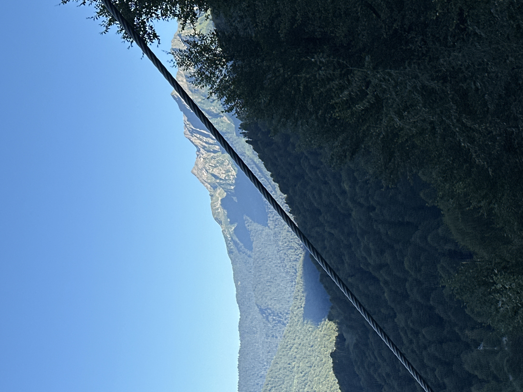 The image shows a view of mountains and trees under a clear blue sky, with a cable extending diagonally across the frame from the lower left to the upper right. The mountains in the background are covered with dense green forests, with some rocky peaks visible at the highest elevations. The cable appears to be part of a ski lift or similar transportation system. The lighting suggests it is daytime, likely morning or afternoon, with the sun casting shadows on the mountain slopes. The scene is natural and scenic, likely taken from a vantage point within or near a mountainous resort area such as Estosadok, Russia, known for its ski resorts and natural beauty. There are no people or specific objects clearly identifiable besides the natural elements and the cable.