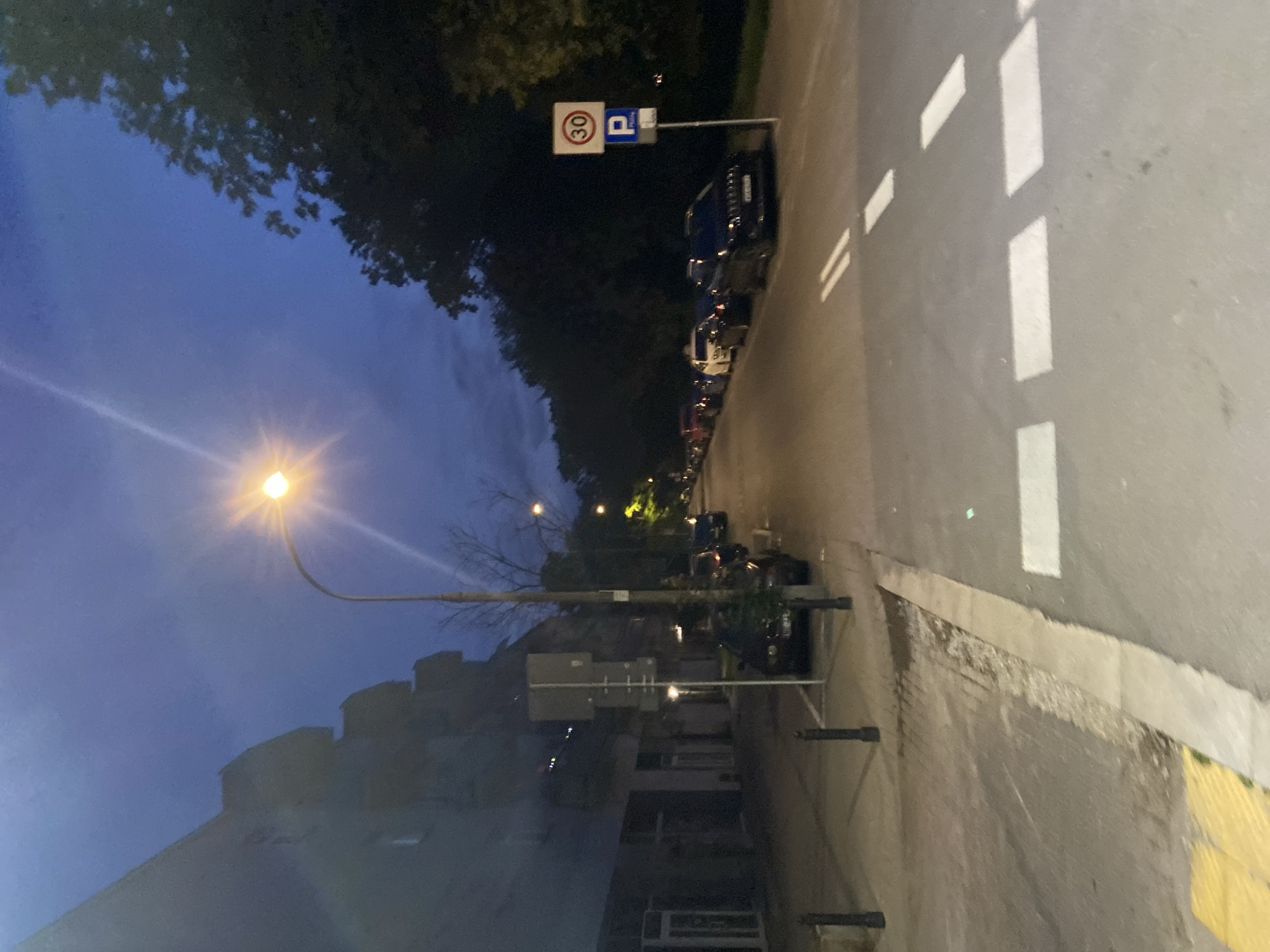 The image shows a street scene in Warsaw, Poland, at dusk. A street lamp illuminates the scene, casting a bright light in the sky. Buildings line the left side of the street, with shops on the ground floor. Several cars are parked along the right side of the street, and a sign indicating a 30 km/h speed limit and parking is visible. Trees and foliage create a dark backdrop on the right side of the image. The street has white markings, indicating lanes and a pedestrian crossing. The overall atmosphere is calm and quiet, typical of a city street at this time of day.