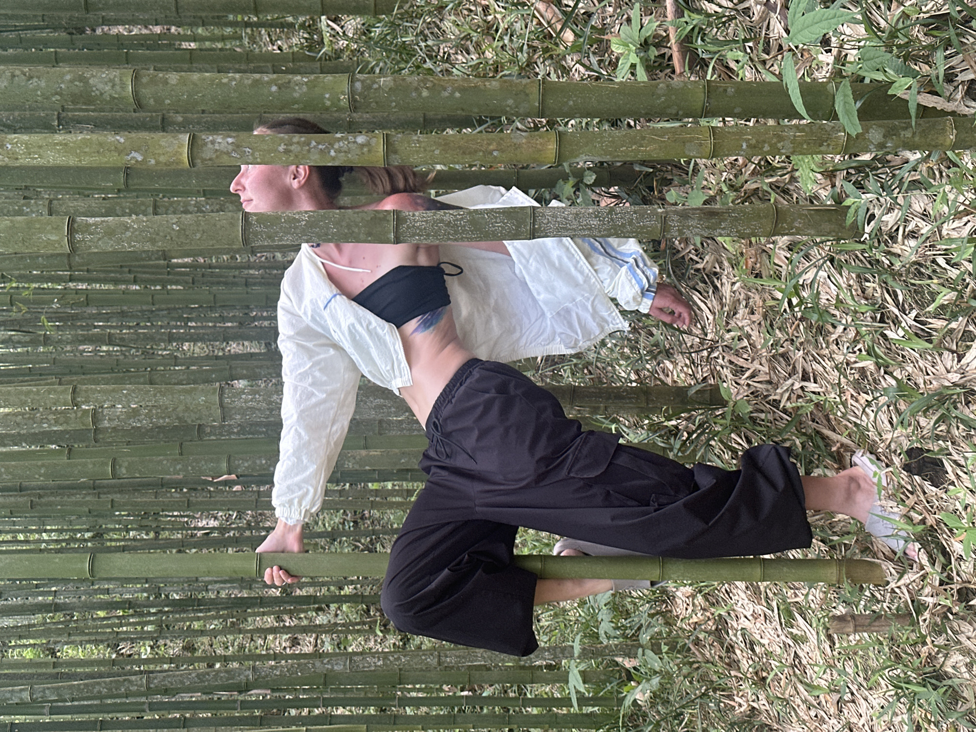 The image shows a woman lying on bamboo in a bamboo forest. She is wearing a black sports bra, a white jacket, and black pants. The background is a dense bamboo forest, consistent with the natural environment in Ngã Ba Kim, Vietnam. The woman appears to be posing or relaxing amongst the bamboo stalks.