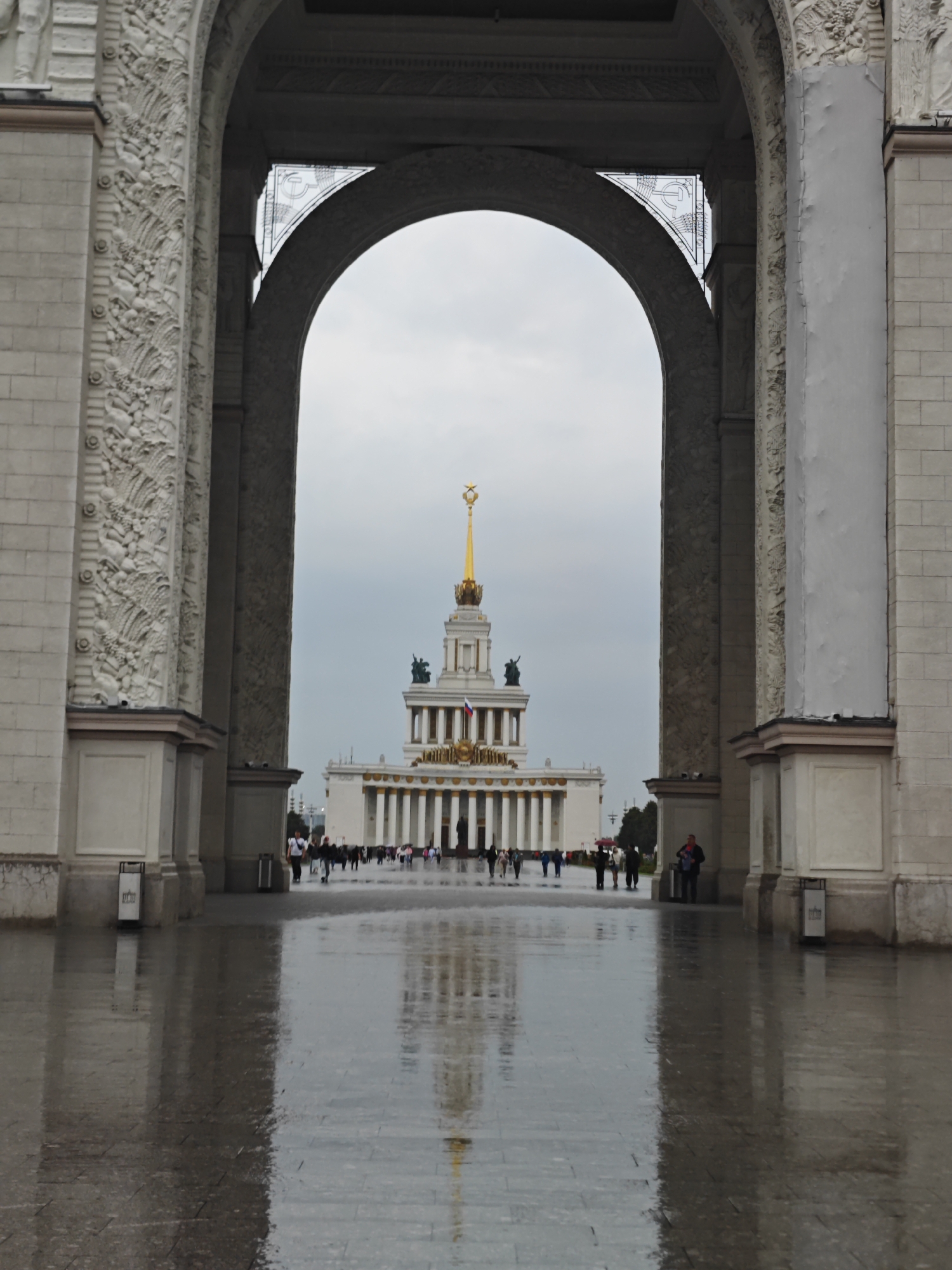 The image shows an archway framing a view of the Main Pavilion at VDNKh (Exhibition of Achievements of National Economy) in Moscow, Russia. The archway is made of stone and decorated with relief sculptures. The ground in front of the archway is wet and reflects the surrounding architecture, suggesting it has recently rained. A number of people are walking in the distance towards the pavilion, which is a grand white building with a tall golden spire topped by a star. The sky is overcast.
