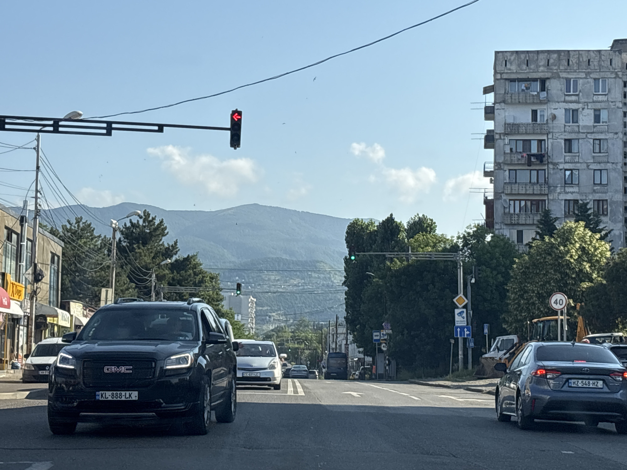 The image depicts a street scene in Tbilisi, Georgia, under a clear blue sky with scattered clouds. The foreground features a black GMC SUV driving on a paved road. The SUV has its headlights on and a license plate reading "KL-888-LK". Several other vehicles are visible, including a white car to the left, a white minivan further ahead, and a dark sedan with a license plate "HZ-548-HZ" to the right.
A traffic light with a red arrow pointing left is suspended above the road. In the background, an old, weathered apartment building with balconies and visible laundry can be seen. Trees line the sides of the street, and further in the distance, rolling mountains are visible. A yellow construction vehicle, possibly an excavator, is parked to the right of the road. A speed limit sign indicating "40" is also present. The overall environment suggests a moderately busy urban setting with elements of residential and commercial infrastructure.