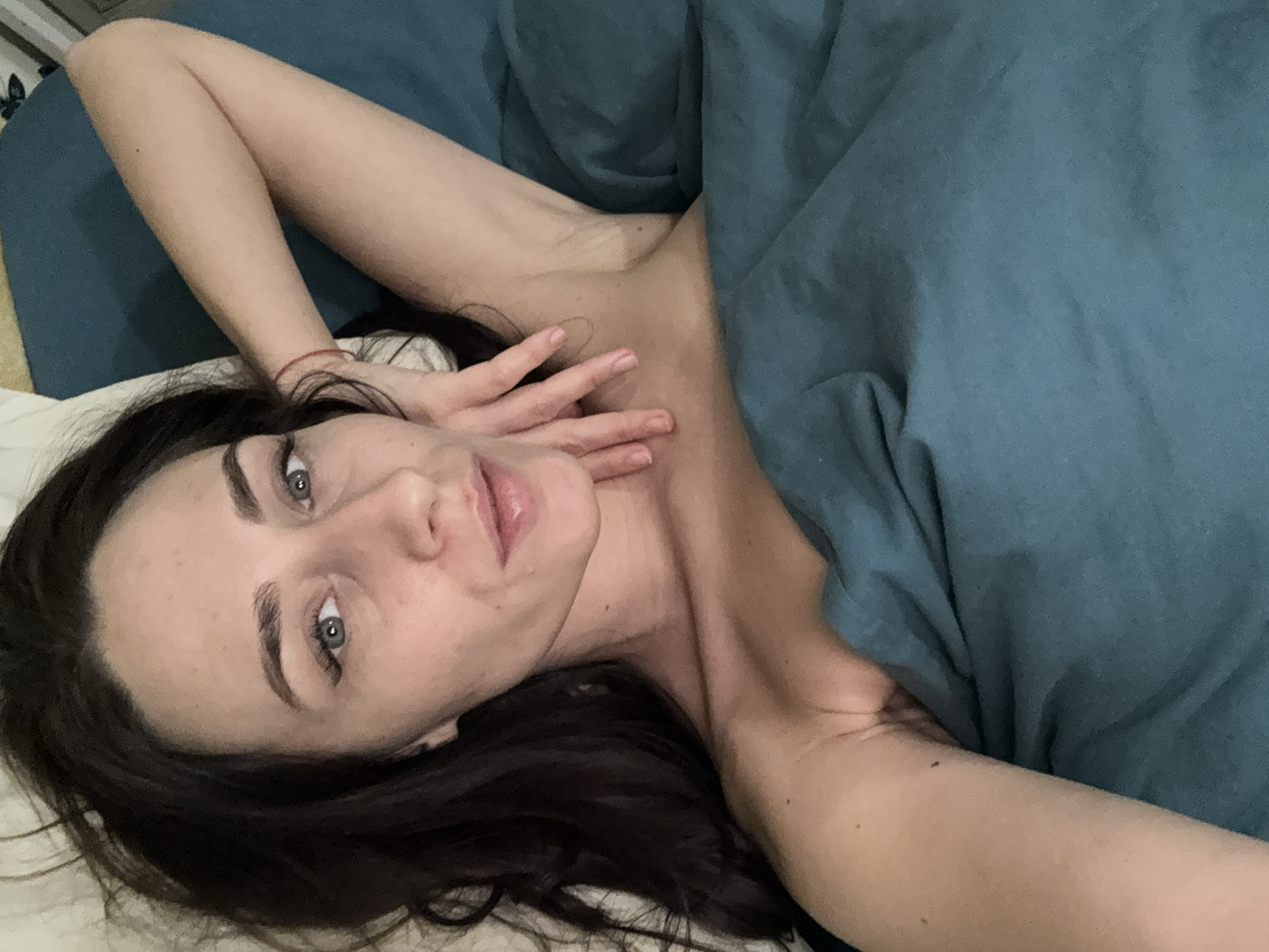 A woman with dark hair and light eyes lies on a bed covered in a blue sheet and white pillow. She is positioned at an angle, with her head resting on the pillow and her hand gently touching her neck. Her gaze is directed toward the camera. The background is out of focus, and the location cannot be determined from the image.