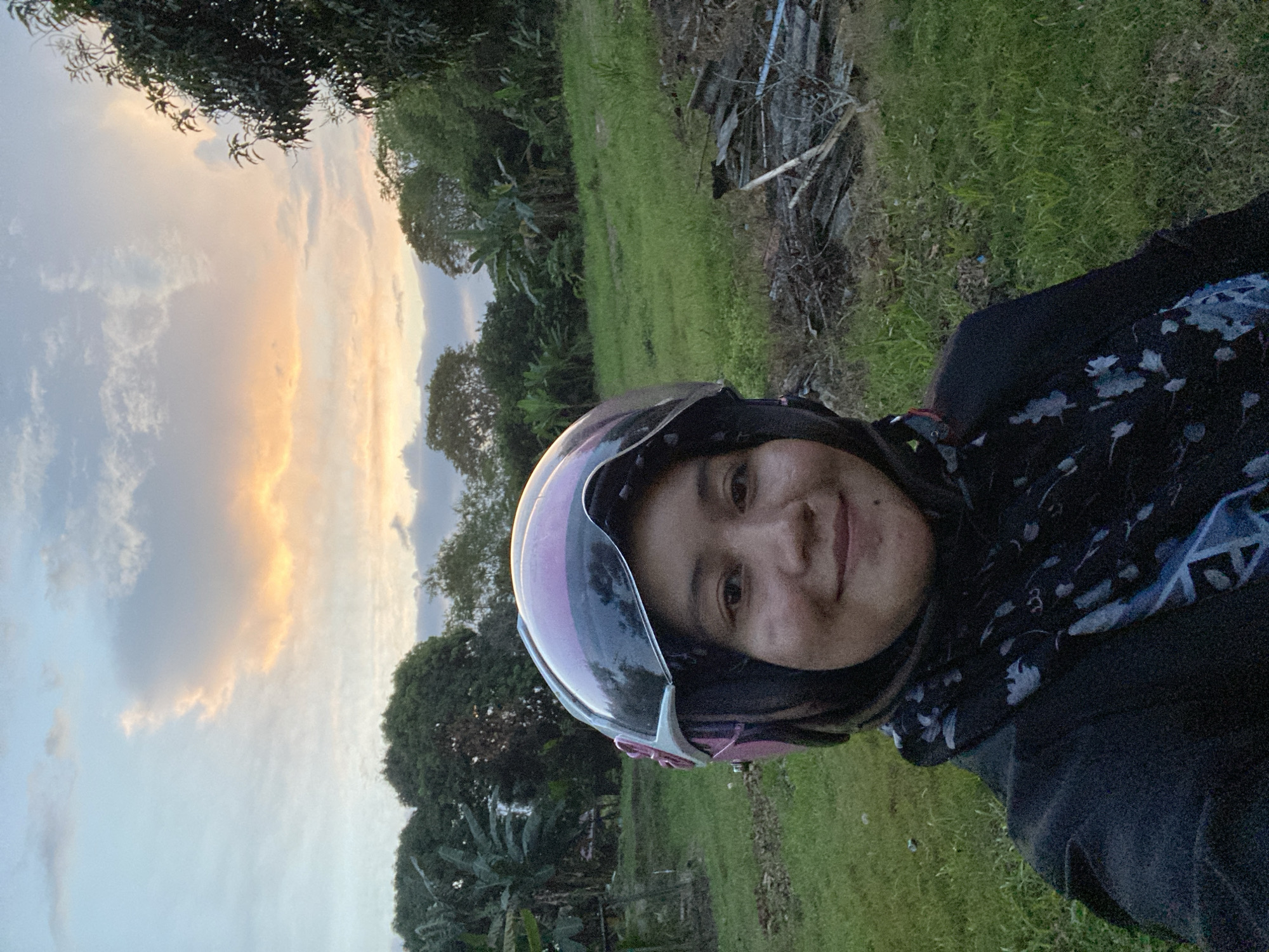 A person wearing a pink and white helmet and a black hijab is pictured outdoors in Banjarbaru, Indonesia. The person is looking at the camera and smiling. The background consists of a grassy field with trees and a cloudy sky with hints of sunset colors. There is also some debris and discarded materials in the foreground.