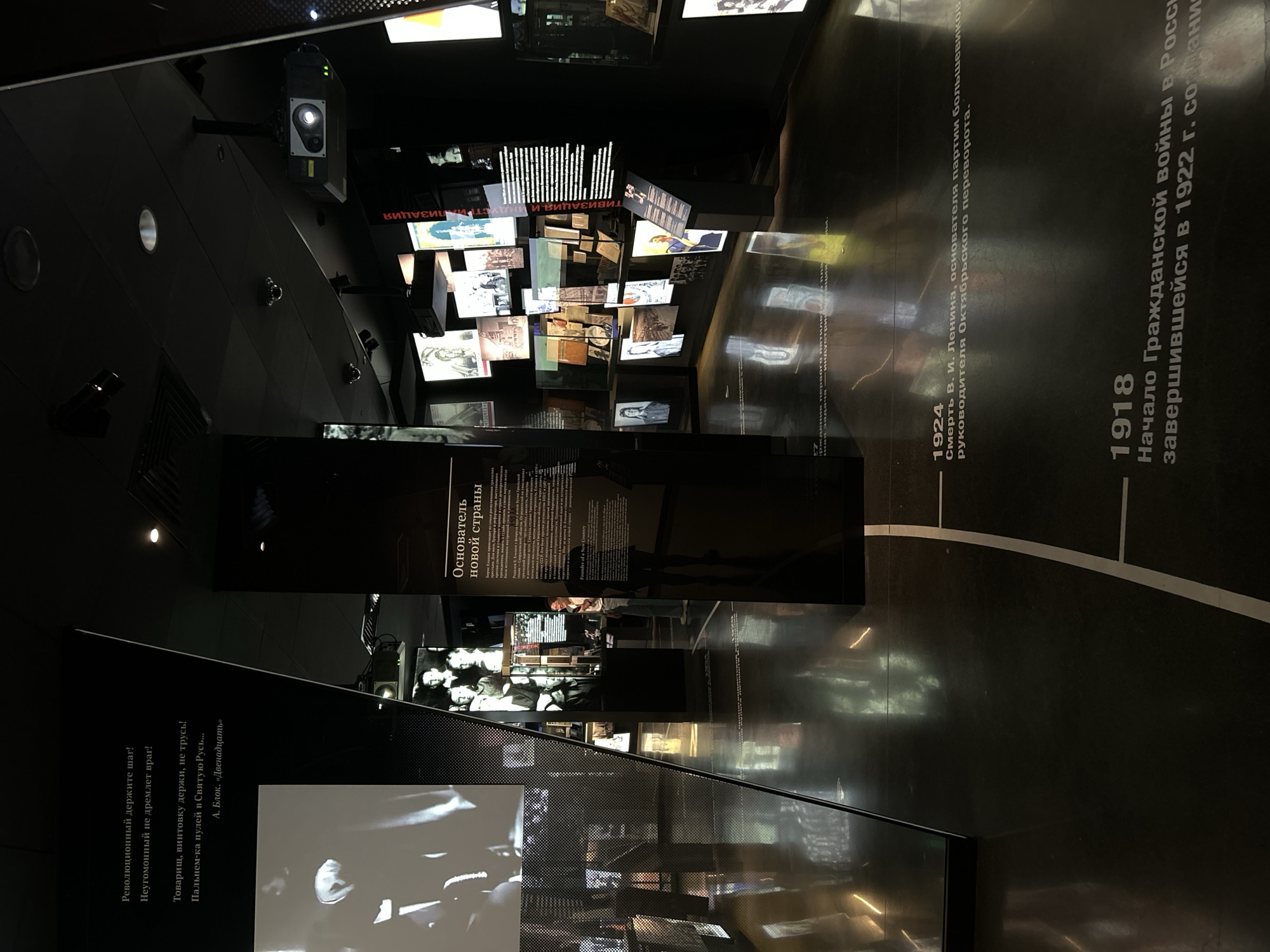 The image depicts an interior exhibition space, likely a museum, with a dark, modern aesthetic. Dominant elements include reflective black walls and floors, creating a sense of depth and showcasing numerous displays. The exhibition features text panels with Russian inscriptions, historical photographs and posters displayed on walls and in display cases, and what appear to be multimedia screens or projections. A projector is visible mounted on the ceiling.
Specifically, the text panels and displayed materials strongly suggest the exhibition is related to Russian history, particularly the revolutionary period and early Soviet era. One prominent text panel in the center displays "Основатель новой страны" (Founder of a New Country) with accompanying text in Russian, and a translated English version below, identifying Boris Yeltsin. Another text panel in the lower portion of the image lists dates and events, including "1924 Смерть В. И. Ленина" (1924 Death of V.I. Lenin) and "1918 Начало Гражданской войны в России" (1918 Beginning of the Civil War in Russia).
The overall atmosphere is serious and educational, with spotlights illuminating specific displays. No visitors are clearly visible interacting with the exhibits, though a figure of a person is partially seen in the mid-right of the image, standing near a display. The architectural style and the nature of the exhibits suggest a significant historical institution. Given the context of the exhibited content, this exhibition is likely located in Russia, and potentially in a city with strong connections to the events depicted. However, the image itself does not provide explicit visual cues to pinpoint Yekaterinburg.