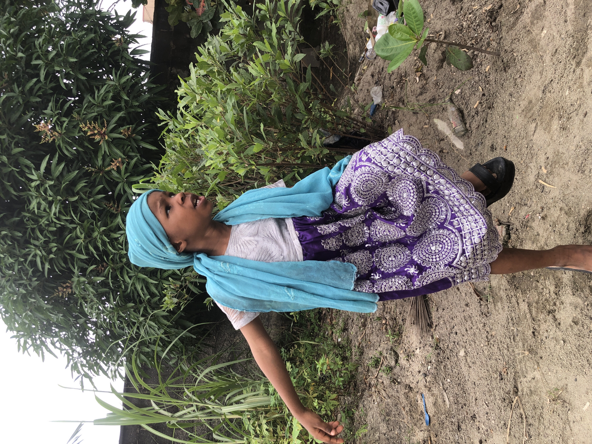 The image captures a young person, appearing to be a child, standing outdoors in what appears to be a residential area in Ado, Nigeria. The individual is wearing a light grey t-shirt, a vibrant purple skirt with intricate white circular patterns and an embellished hem, and a light blue headscarf and matching scarf draped around their shoulders. Their left arm is extended outwards, and their right leg is lifted, suggesting movement or a posed stance.
The environment is somewhat untidy, with dry, sandy ground. Several plants are visible, including tall green stalks resembling sugarcane on the left, a dense green shrubbery in the middle ground, and a small sapling on the right. A low, dark-colored wall or fence is present behind the vegetation on the left. Scattered on the ground are various items, including a blue plastic spoon, a broken glass bottle, and plastic debris like bottles and bags, indicating litter. In the background, the foliage of a large tree with some flowering branches is prominent. The sky appears overcast. The scene suggests an outdoor space within a community, possibly a backyard or an undeveloped plot of land.