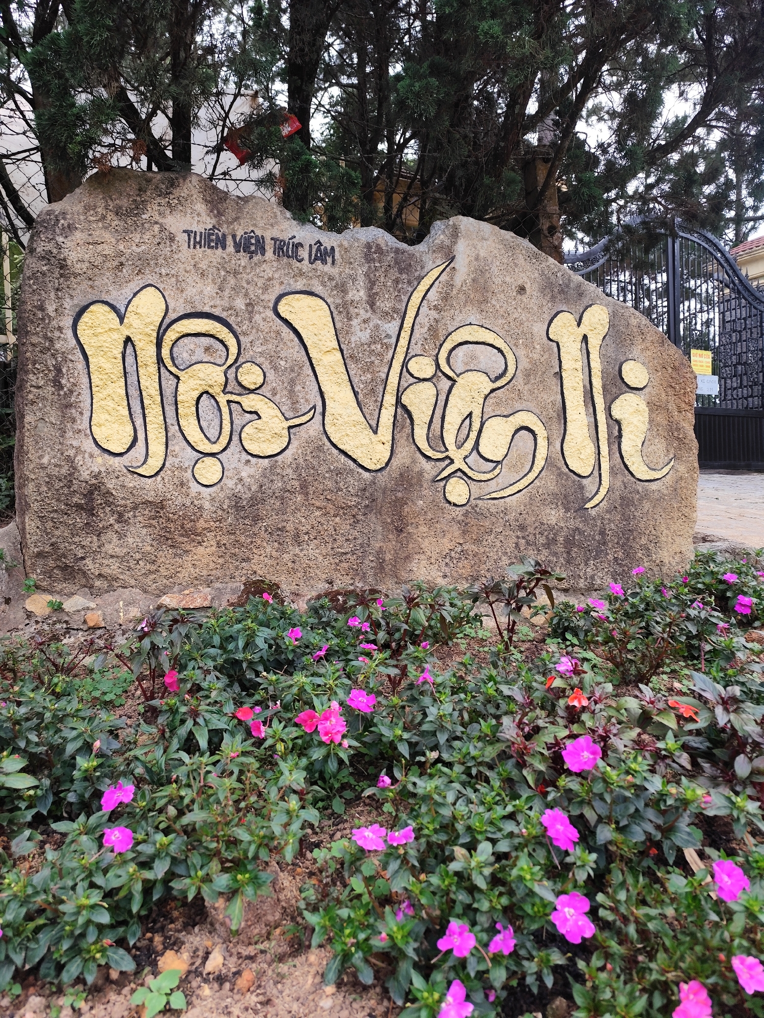 The image shows a stone sign in front of a building with a gate. The sign is inscribed with the words "Thien Vien Truc Lam" above and "Ho Voi Li" below in yellow lettering. The base of the sign is surrounded by a flower bed with pink and red flowers. Behind the sign are trees and a black wrought-iron gate. The location is likely Da Lat, Vietnam, based on the Vietnamese text on the sign.