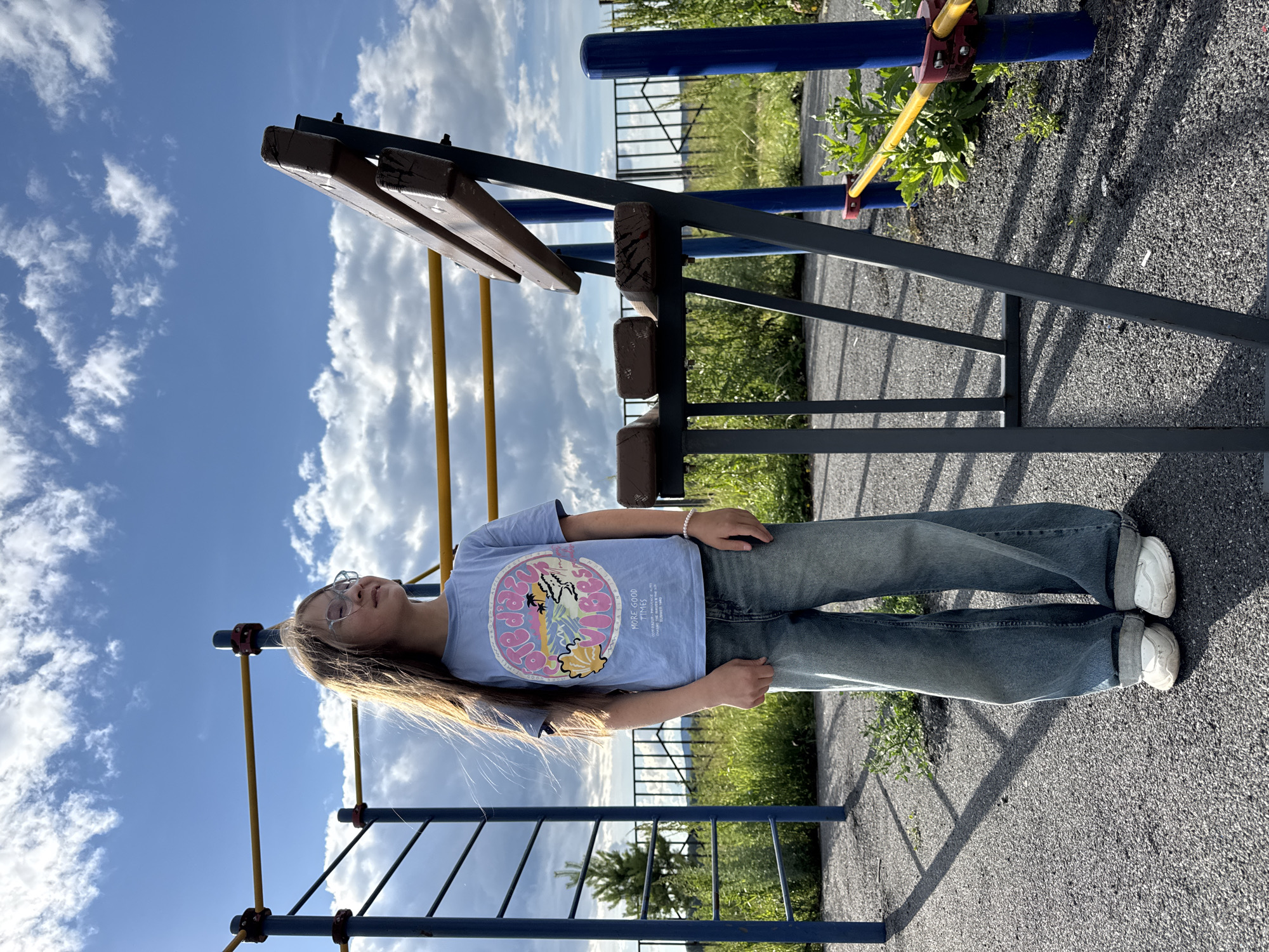 This media file shows a young person standing on a playground in Mursalimkino, Russia. The person is wearing a light blue t-shirt with a circular graphic on it, blue jeans, and white sneakers. They have long, light brown hair and are wearing glasses. The graphic on the t-shirt appears to depict a wave, a beach, and a sun, with text that is partially obscured but includes "MORE GOOD TIMES."
The playground equipment is visible, including metal bars and wooden beams, painted in blue, yellow, and dark brown. The ground is covered in dark gravel. In the background, there is green grass and a metal fence. The sky is blue with fluffy white clouds, suggesting it is daytime. The lighting indicates it is likely midday or afternoon. The overall mood seems relaxed and casual, consistent with a day spent outdoors.