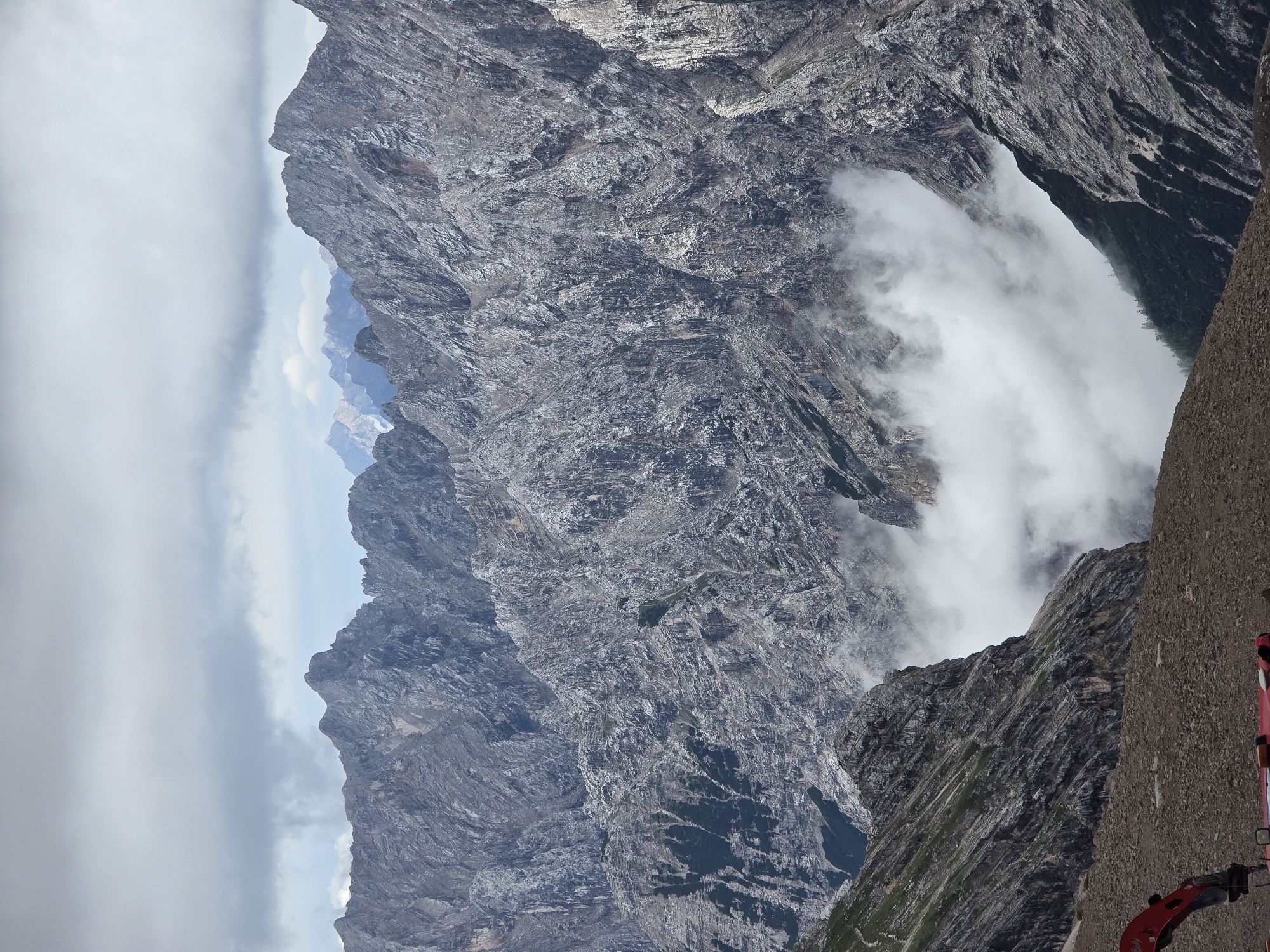 The image shows a majestic mountain landscape under a cloudy sky, likely in the Bavarian Alps near Garmisch-Partenkirchen, Germany. The foreground features a rocky, barren terrain with a small section of red construction equipment, possibly a snow blower or similar machinery, visible at the bottom left. Beyond this, steep, rugged mountains dominate the scene, with sheer rock faces characterized by deep crevices and weathered textures. Wisps of white clouds or fog are present in the valleys and lower elevations between the mountain peaks, adding a sense of depth and atmosphere. In the distance, more mountain ranges are visible, some appearing lighter in color and possibly covered in snow, hinting at higher altitudes. The overall impression is one of a dramatic and imposing natural environment, with a touch of human presence represented by the construction equipment in the foreground.