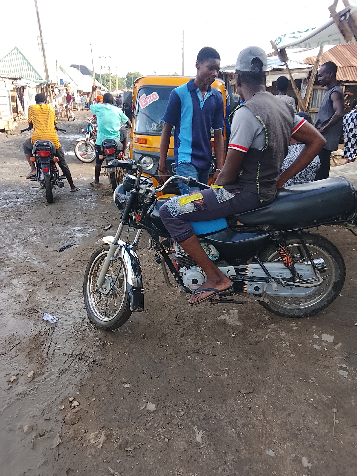 The image depicts a street scene in Abuja, Nigeria, characterized by a muddy ground and a collection of motorcycles and a yellow tricycle (a "keke"). Several people are present, some standing near the vehicles and others on motorcycles. One man is seated on a motorcycle, seemingly engaged in conversation with another individual standing next to him. Other figures are visible in the background, suggesting a bustling environment. The architecture consists of basic structures, some with corrugated metal roofs.