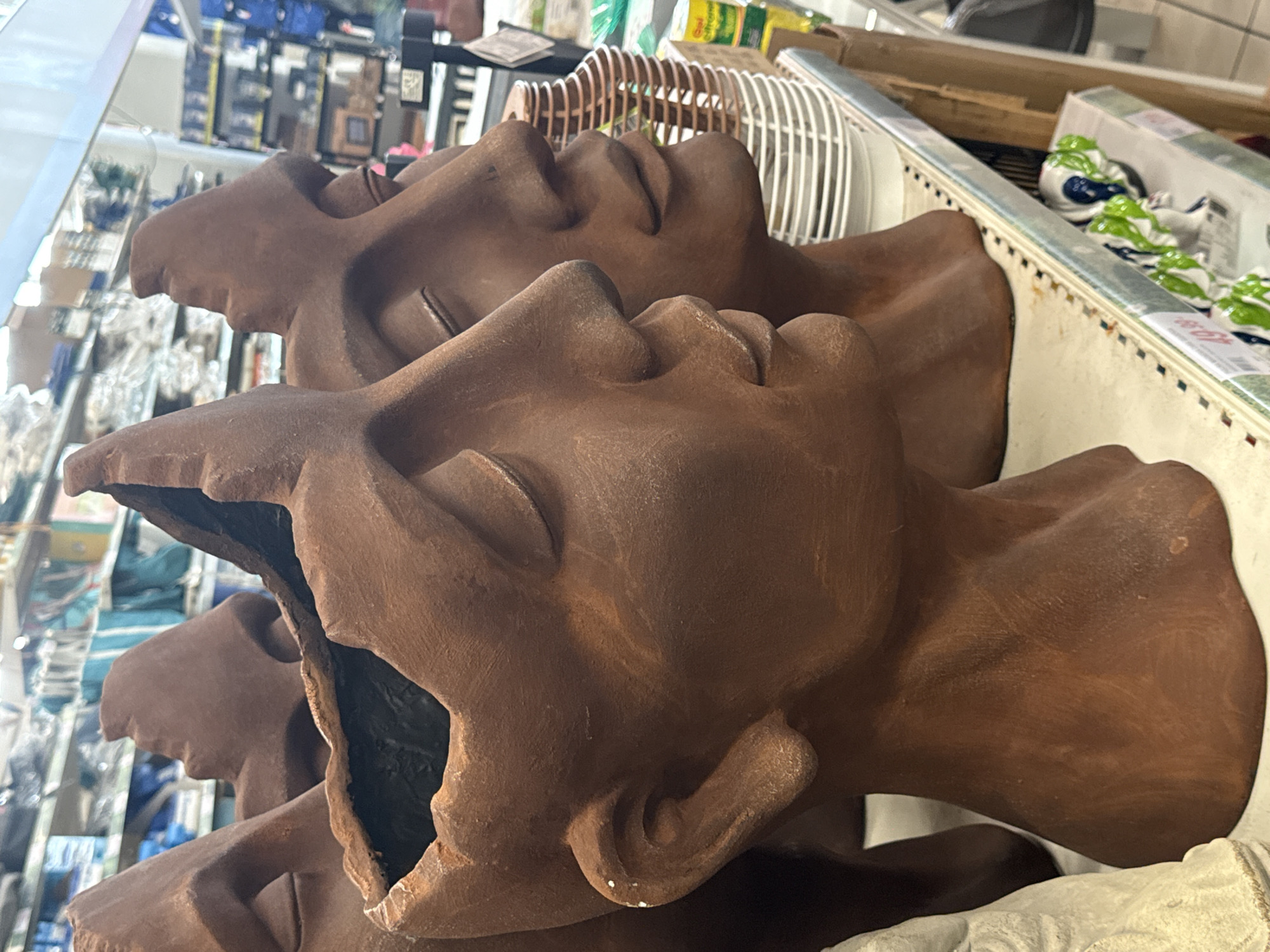 The image shows a close-up view of three brown, head-shaped planters arranged on a shelf in what appears to be a store. The planters are made of a rough, clay-like material and have an opening at the top, revealing a dark interior. The planter in the foreground is the most prominent, showing the profile of a face with closed eyes and a slightly open mouth. To the right of this planter are two other similar planters, positioned slightly behind and to the side. The background is blurred but suggests a retail environment with shelves filled with various items, including bags, boxes, and other decorative objects. Some price tags are visible on the shelves. The lighting is typical of an indoor retail space.