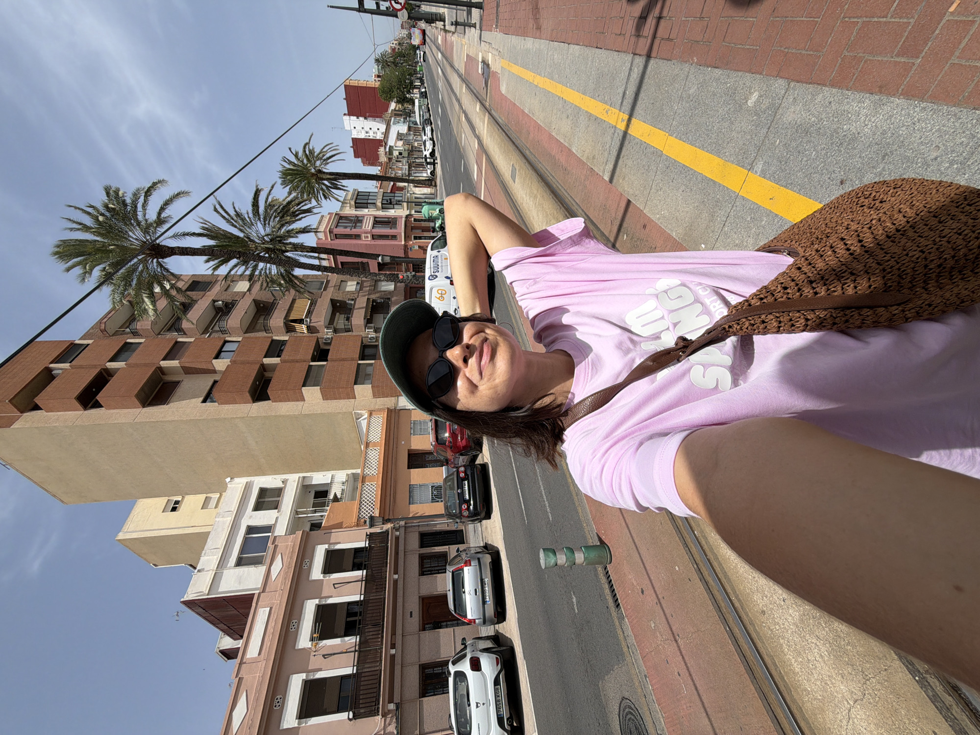 This media file is a selfie of a person taking a picture of themselves. The person is wearing a pink t-shirt, sunglasses, and a dark green cap. They have a brown woven bag on their shoulder. The scene appears to be outdoors, possibly on a sidewalk next to a street with tram tracks. Buildings with palm trees can be seen in the background. The lighting suggests it is daytime, and the weather appears to be sunny. The person's facial expression is relaxed and pleasant, indicating they are enjoying themselves. The orientation of the image is sideways. The visible text on the t-shirt appears to be a white graphic with some lettering.
