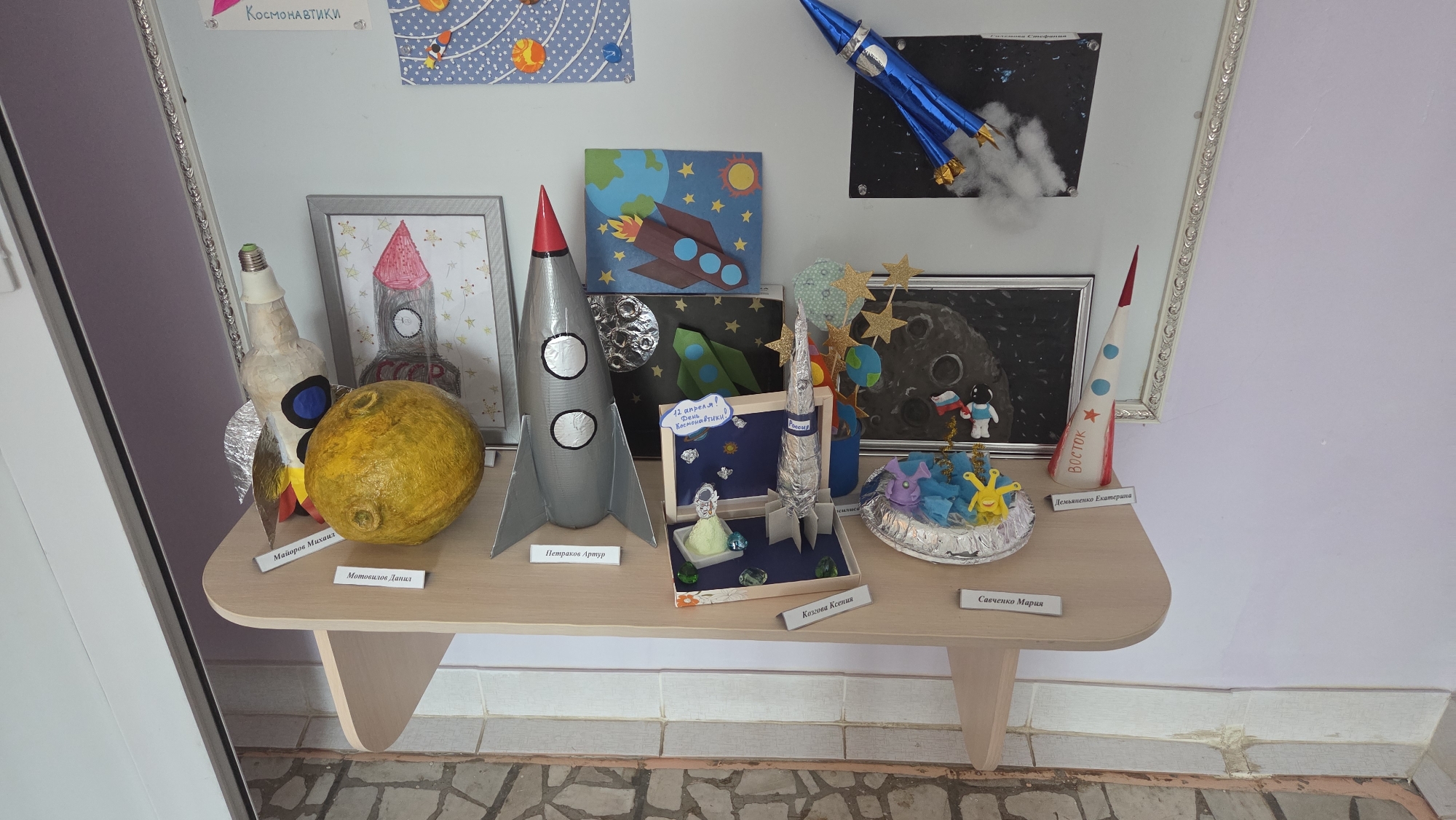 The image captures a display of space-themed children's artwork, likely from a school or library in Chaikovsky, Russia. The creations are displayed on a light-brown wooden table set against a wall. The wall has a section painted light purple above the table, and a white section above that. The artwork includes models of rockets, the moon, drawings of rockets and planets, and depictions of astronauts. The artwork includes the labels "Майоров Михаил", "Мотовилов Данил", "Петраков Артур", "Козгова Ксения", "Савченко Мария", and "Демьяненко Екатерина," which are presumably the names of the young artists. A sign with "12 апреля! День Космонавтики." is among the art. The items are arranged in a row, showing a variety of materials and artistic styles, suggesting a school project or competition related to space exploration. The floor is covered with grey and white square tiles.