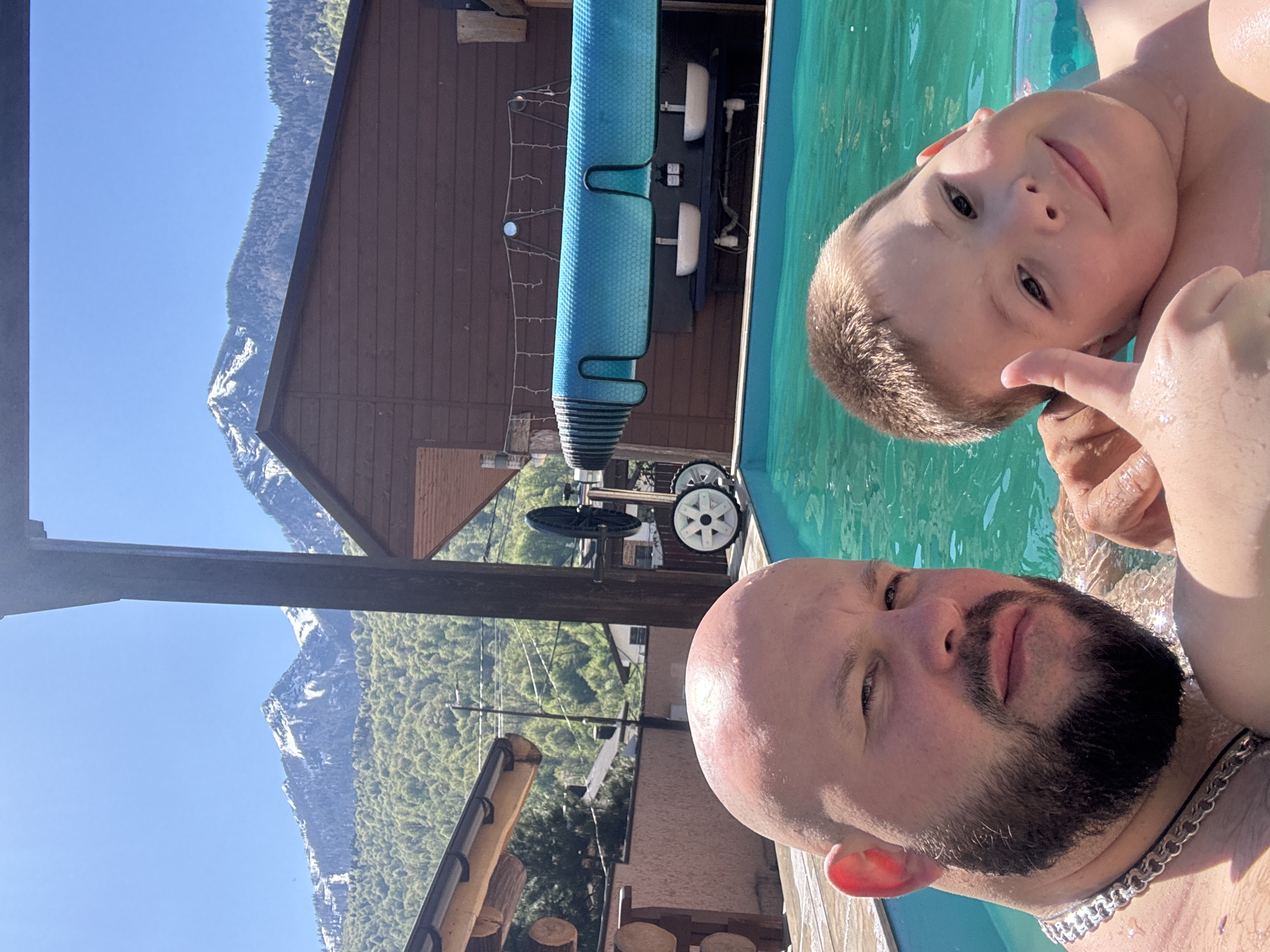 A bald man with a black beard is in a swimming pool with a young boy, presumably his son, in the city of Krasnaya Polyana, Russia. The boy is giving a thumbs up. Behind them, there is a house or building with a wooden exterior and a pool cover. In the background, snow-capped mountains are visible under a clear blue sky.