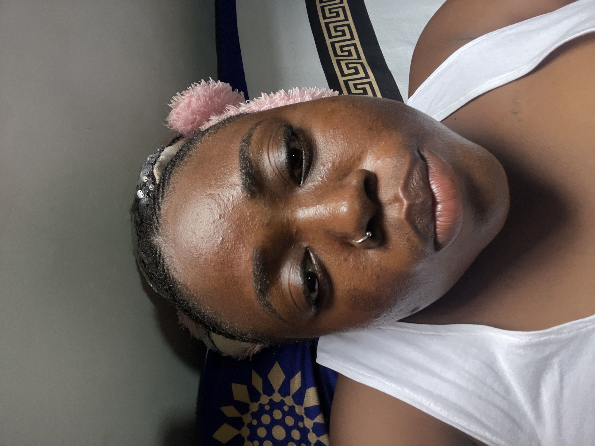 A close-up, low-angle selfie shows a Black woman lying down, possibly on a bed, with a blue and gold patterned pillow behind her left shoulder and a black and gold striped blanket or pillowcase behind her right shoulder. She is wearing a white tank top and a pink fluffy headband with sequins. Her hair is braided and pulled back. She has a silver hoop nose ring. Her skin appears dewy and she is looking down and slightly to the left with a neutral expression. The image quality is slightly grainy, with visible skin texture and light reflections. The setting appears to be indoors, with a plain gray wall visible in the background. There is no indication of the city of Abuja, Nigeria, in the image.