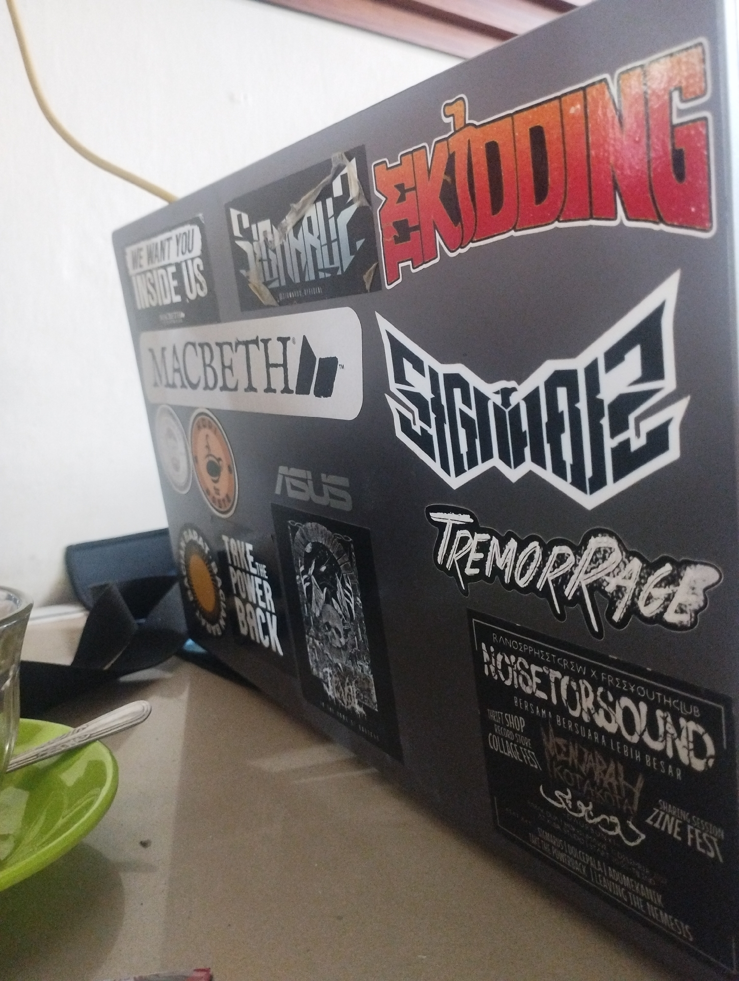 The image shows an Asus laptop covered in various stickers. These stickers include phrases like "WE WANT YOU INSIDE US", "MACBETH", "TAKE THE POWER BACK", "TREMORRAGE", and "NOISETORSOUND". The laptop also has a "SIGNAL2" sticker and "SKIDDING" in large red letters. A green saucer with a spoon and a glass are visible on a tan-colored surface in the foreground. The location, Lhokseumawe, Indonesia, is not visually discernible from the image.