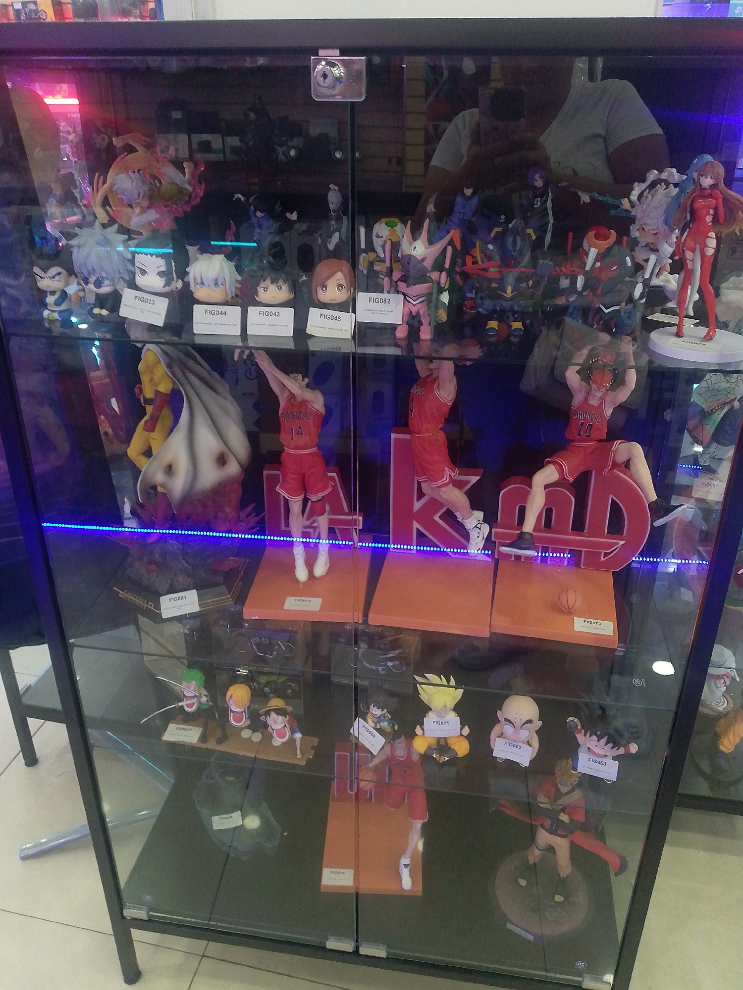 The image depicts a black-framed glass display cabinet filled with anime and manga figurines. The figurines are arranged on multiple shelves illuminated by a blue LED strip.
On the top shelf are various figurines, including smaller head figures with identification tags, and larger figures, including one in a red outfit.
The middle shelf features basketball-themed figurines, some with "SHOHOKU 14" on their jerseys, along with a figure with a large white cape.
The bottom shelf contains other anime figurines, including characters from "Dragon Ball" and "One Piece."
The reflection in the glass reveals a person holding a mobile phone, likely taking the photograph.
The context of the location, "city of Caracas, Venezuela," is not visually discernible within the image itself.