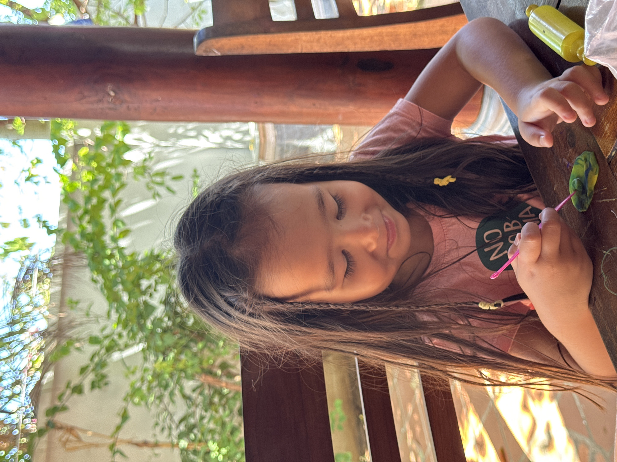 A young girl is seated at a wooden table outside, possibly on a patio, indicated by the presence of greenery and natural light. She has long brown hair with small braids along the side. She is wearing a pink t-shirt with a dark circular design. The girl is focused on working with green and blue clay on the table, using a pink tool. A yellow container is visible on the table. The scene suggests a leisure activity, possibly at a residence or establishment in Mũi Né, Vietnam, though specific landmarks are not apparent to confirm this.