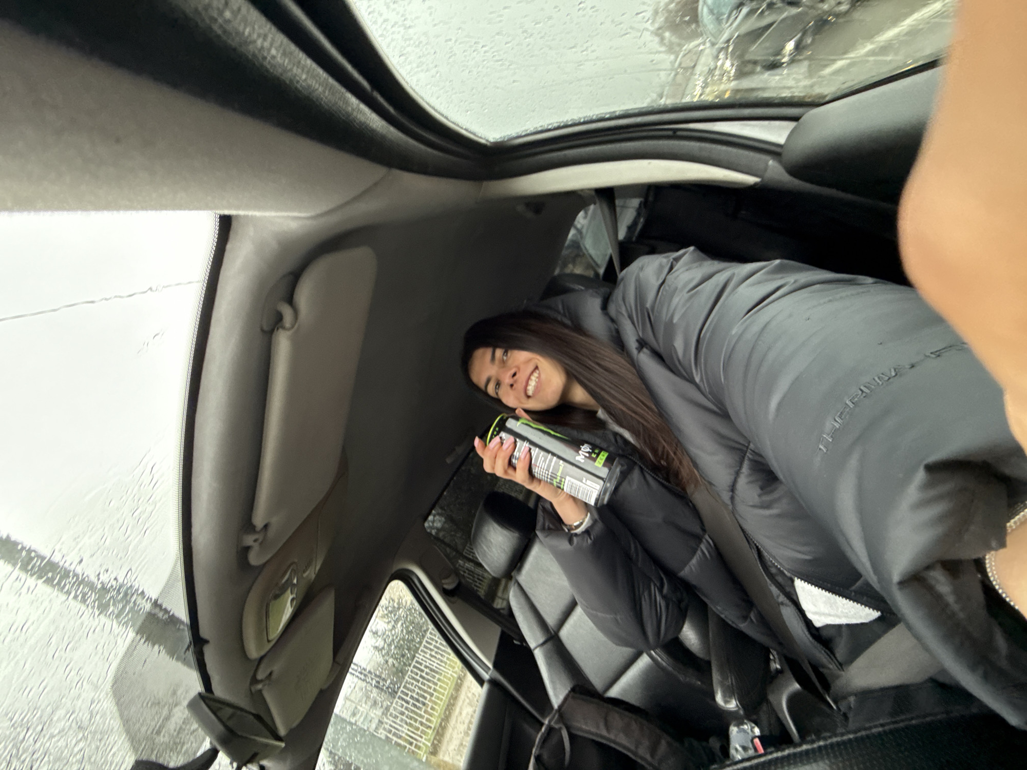 A female individual is depicted in a selfie-style photograph taken from the driver's seat of a vehicle in Sofia, Bulgaria, during rainy conditions. The subject, with long dark hair and a light complexion, is smiling directly at the camera. She is wearing a dark, possibly black, puffy winter jacket over a lighter garment, with a white zipper detail visible. Her left hand holds a black and green Monster Energy Zero Sugar can, displaying pink nail polish on her fingers and a silver-colored bracelet on her wrist. The car interior features black leather seats and dark-colored trim. A dark backpack is visible on the unoccupied front passenger seat. Through the rain-streaked windshield and side windows, a blurred exterior scene includes a white metal fence, dark vegetation, and indistinct vehicles, consistent with an urban or suburban street environment. Sun visors and a rearview mirror are visible on the car's interior ceiling.