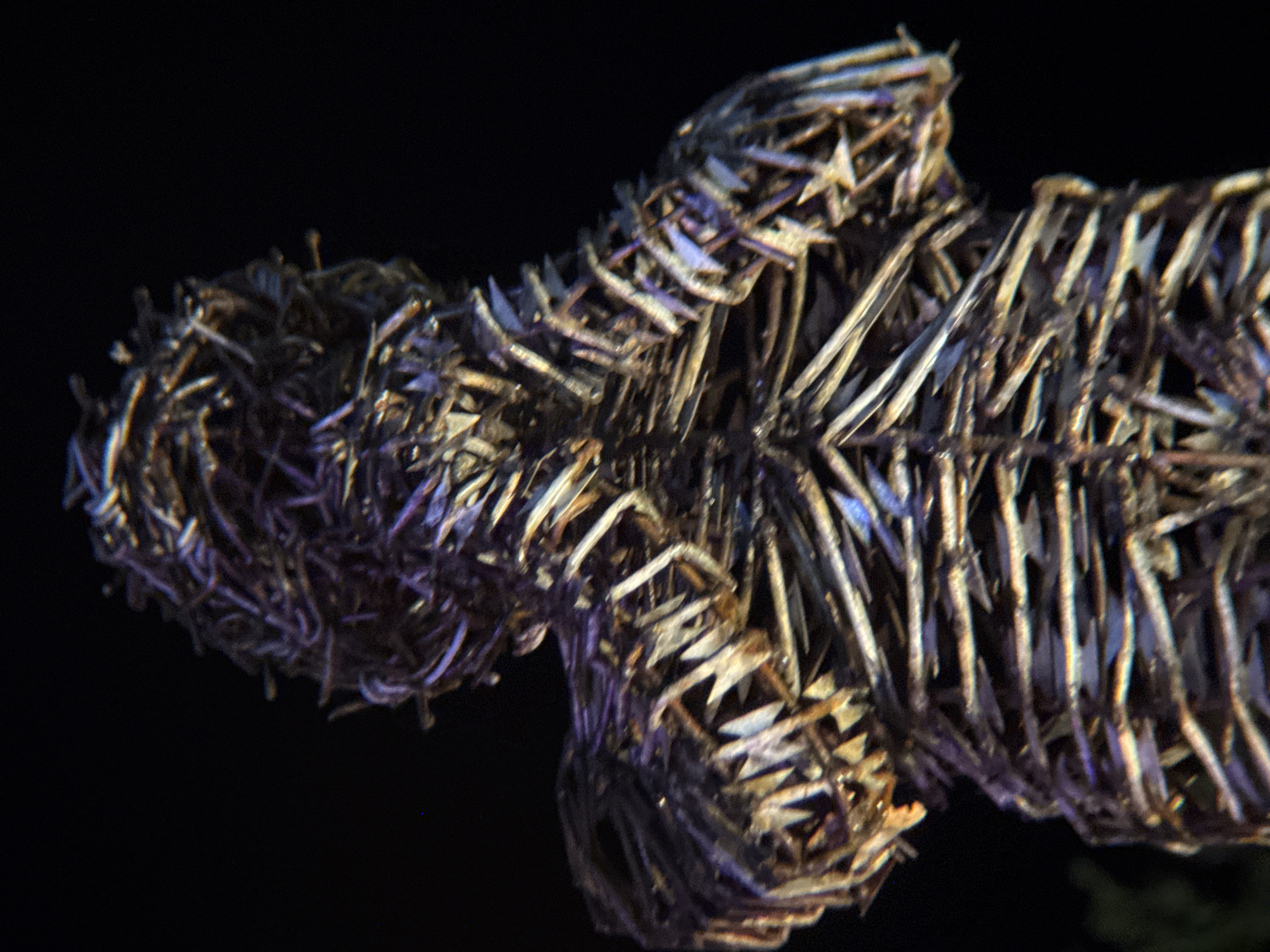 The image shows a close-up of a woven figure, possibly a statue or effigy, against a dark background. The figure appears to be made of thin, fibrous material, woven tightly together to form a humanoid shape. The lighting highlights the texture of the weave, creating shadows and catching iridescent colors that suggest a metallic or reflective quality to the material. The figure's head is densely woven, and the arms are bent at the elbows, held close to the chest. The overall impression is abstract and somewhat unsettling due to the dark environment and the texture of the material. There is no discernible location in the city of Thuận Hóa Ward, Vietnam within the image.