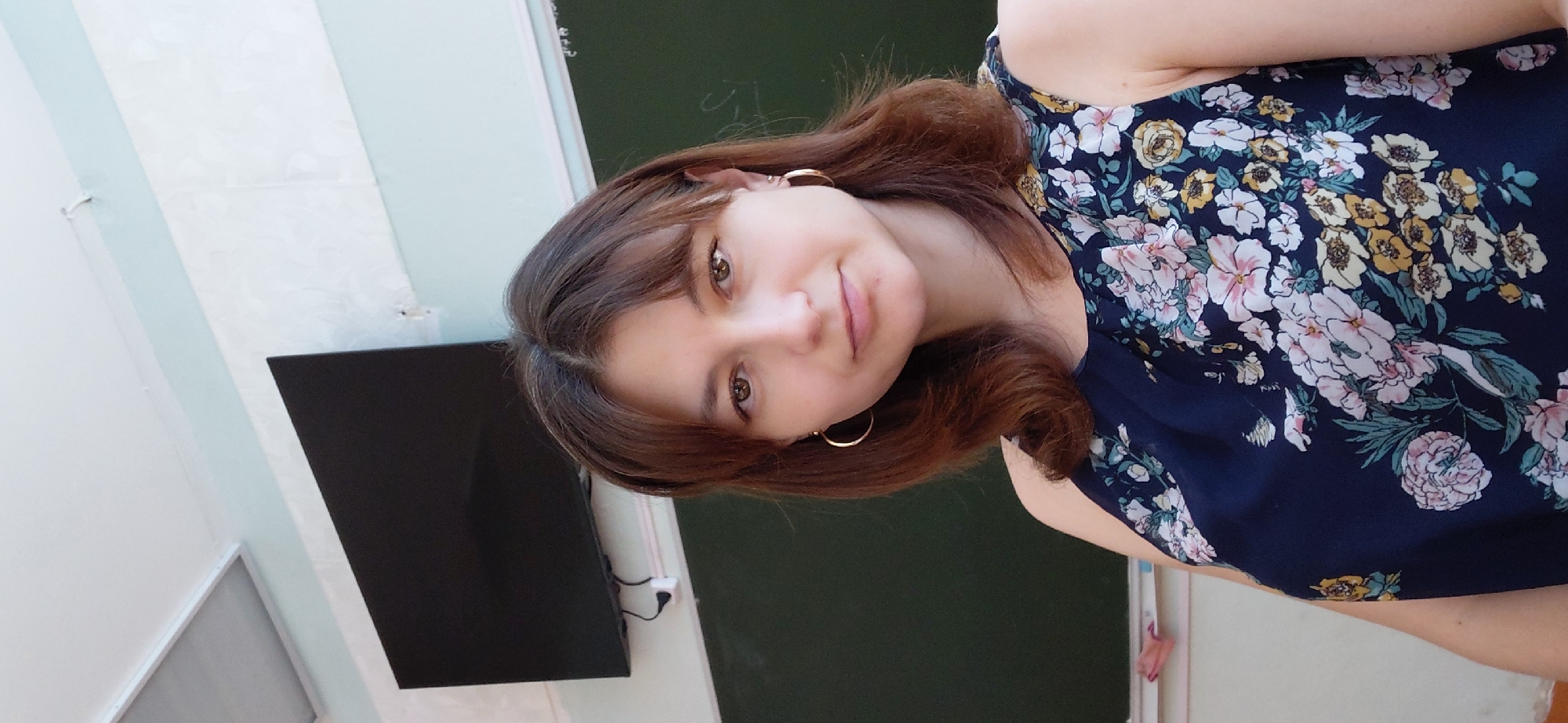 The image shows a young woman with brown hair and brown eyes, wearing a dark blue floral dress and gold hoop earrings. She is positioned in the foreground, looking towards the camera. In the background, a blackboard is visible with some faint writing on it, and a black television screen is mounted on the wall above the blackboard. There is also a fluorescent light fixture on the ceiling. The overall setting appears to be a classroom or a similar educational environment. The scene is static, with no visible action taking place. The lighting is bright, suggesting it is daytime. The location is identified as Svetlogorsk, Russia.
