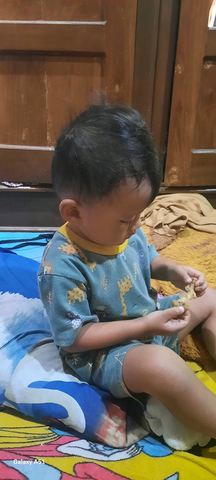 A young child, likely male and of Asian ethnicity, is seated on a colorful patterned bedspread. The child is wearing a short-sleeved t-shirt and shorts, both featuring a blue background with a repeating pattern of giraffes and other cartoonish animals. The child is looking down and holding a small, light-brown object, possibly food, in their hands. The background consists of a wooden cabinet or furniture and a tan blanket. The setting appears to be indoors, in a bedroom. The image was captured in Jatitengah, Indonesia.