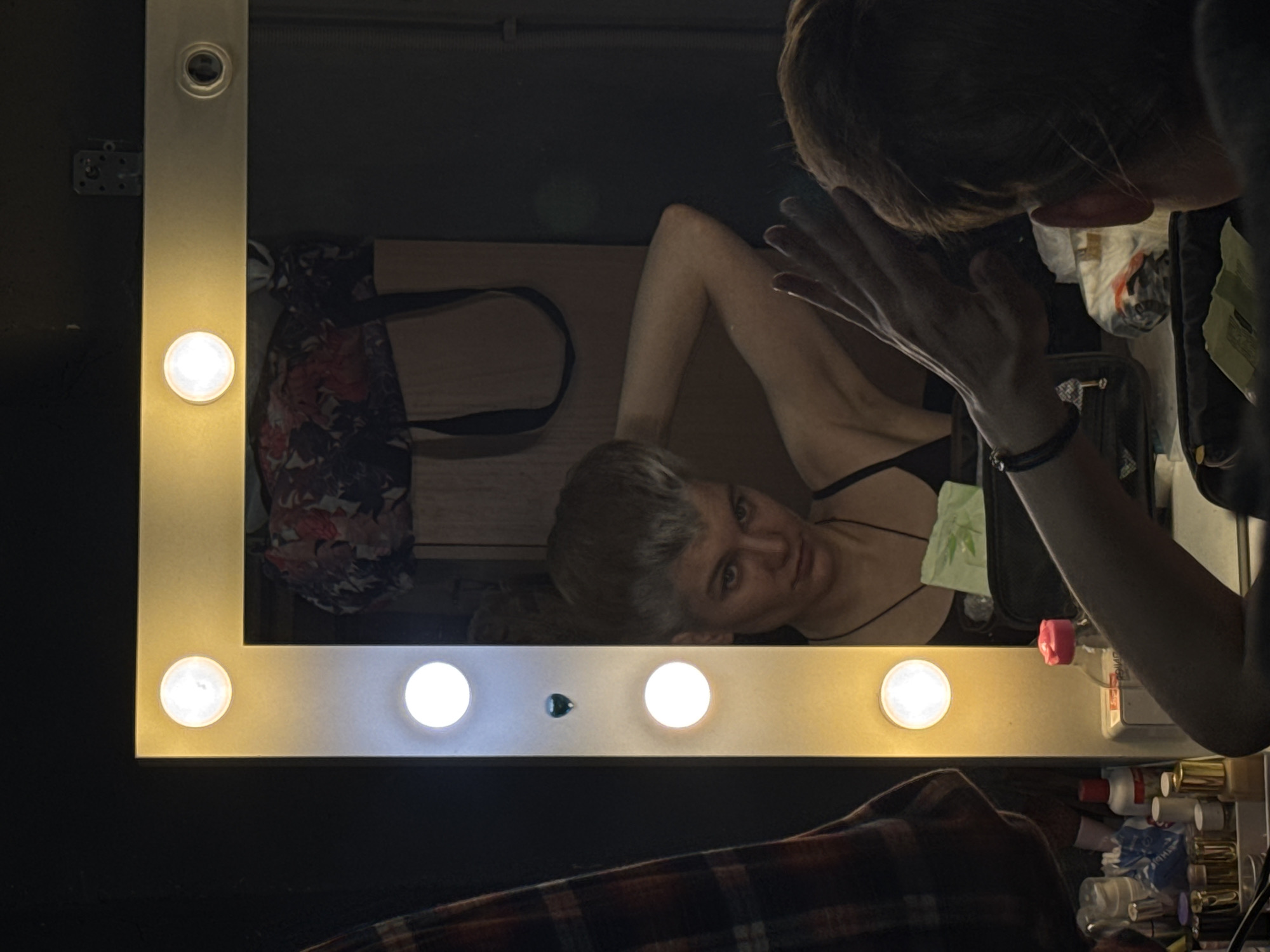 An image captured at a 90-degree clockwise rotation depicts a person interacting with a vanity mirror. The primary subject is a young individual, likely female, whose head and shoulders are visible from behind on the right side of the frame, looking into the large rectangular mirror. Her right arm is raised with the palm of her hand appearing to touch or be close to the mirror's surface.
The mirror's reflection shows the person's face, a neutral to slightly serious expression, and her upper body. She has light-colored hair and is wearing a dark, possibly black, spaghetti strap top. In the reflection, a floral-patterned bag and a wooden surface, likely a vanity table, are visible behind her. Various items are also reflected on the table, including what appears to be a pink-capped plastic water bottle, other cosmetic bottles or containers, and a crumpled green tissue or napkin.
The mirror itself is framed by a light-colored border, likely metal or plastic, featuring several round light bulbs. Some bulbs emit a warm yellow light, while others appear to be a cooler white. On the top left edge of the mirror frame, a small, dark circular opening resembling a camera lens or a small sensor is present.
Outside the mirror's reflection, on the bottom part of the original image (which appears on the left due to rotation), a plaid fabric, possibly a blanket or clothing, is visible. A metal bracket is also attached to the wall near the top left corner of the mirror frame. The overall lighting is provided by the bright vanity bulbs, creating an intimate indoor atmosphere. There is no clearly discernible text.