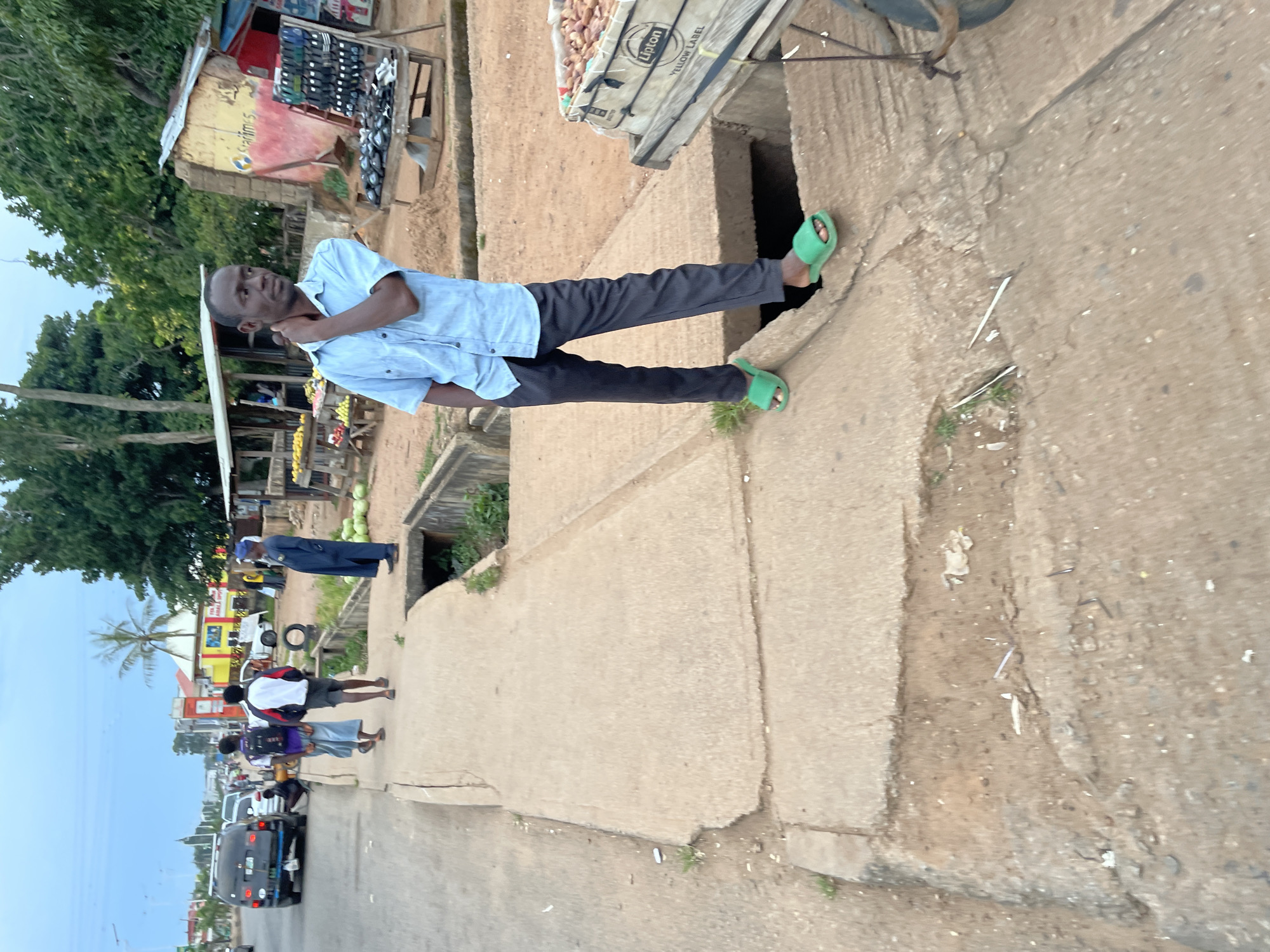 This is a medium shot, eye-level, outdoor, daytime photograph taken in Jos, Nigeria. The scene captures a street with pedestrians, vehicles, market stalls, and a man standing on a sidewalk.
In the foreground, a man in a light blue short-sleeved shirt and dark striped pants stands with his left hand resting on his neck and his right hand in his pocket. He is wearing bright green slide sandals. He is positioned on a concrete sidewalk that appears cracked and uneven, bordering a dirt area with some sparse vegetation. To his right, a market stall on wheels holds produce, identified by a "Lipton Yellow Label" sign.
In the mid-ground, two young men are walking away from the camera on the sidewalk, one wearing a backpack. Further down the sidewalk, another person in a blue robe stands near a small roadside stall. A few green watermelons are visible near the stall.
In the background, a busy street with various vehicles, including a black SUV, is visible. Buildings and trees line the street, suggesting a commercial or residential area within Jos. The sky is overcast, indicating a cloudy day. The overall impression is of an ordinary day in a bustling Nigerian city.