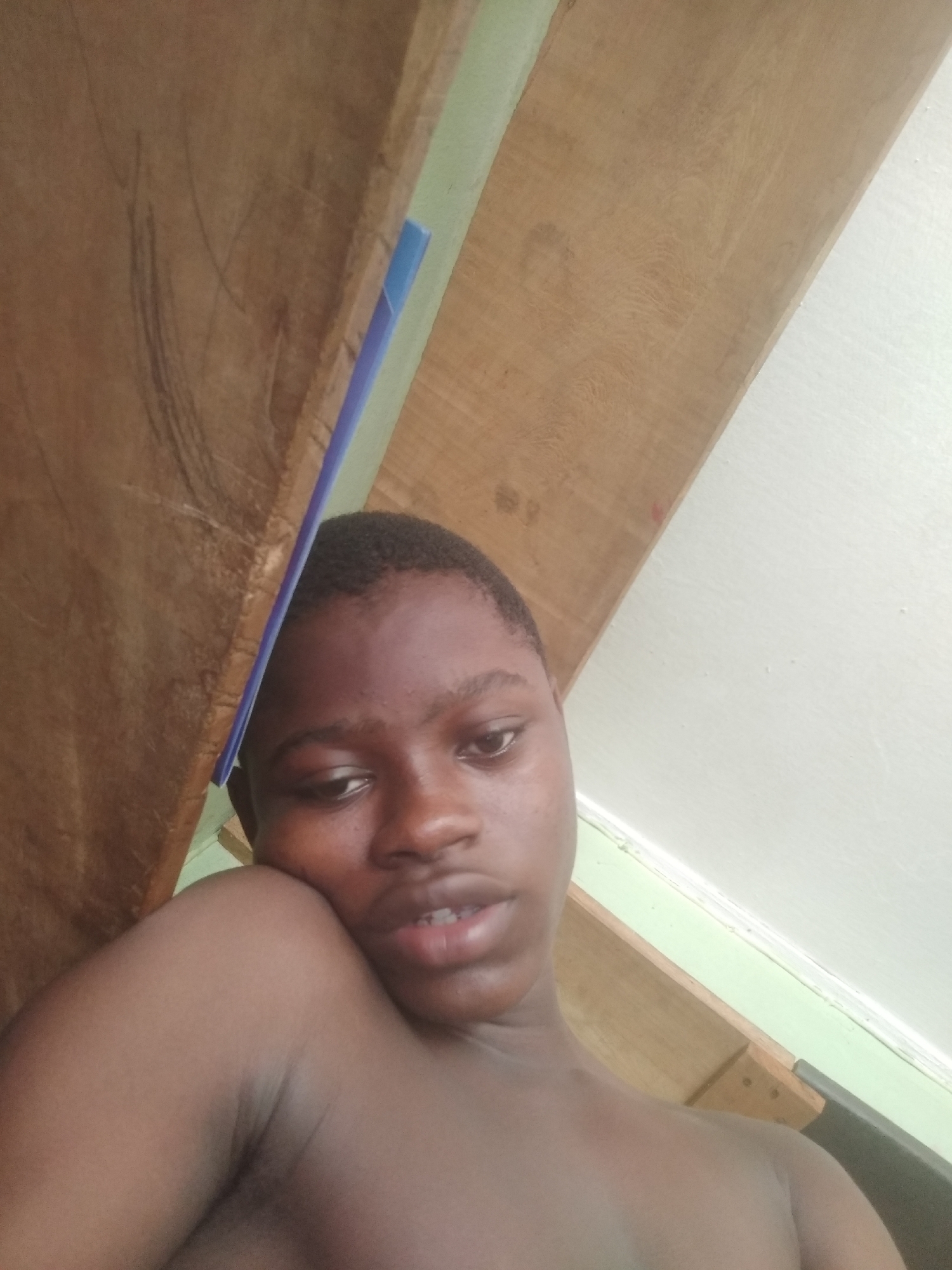 A close-up, low-angle portrait features a young individual with dark skin and short, dark hair, captured in an indoor setting. The person, appearing shirtless, occupies the lower and central part of the frame, looking slightly upwards and to the right with a neutral, relaxed expression and a slightly open mouth.
A large wooden beam or plank dominates the upper and left sections of the image, displaying a rustic texture with visible grain and some dark marks. A thin blue object is partially visible, tucked behind this wooden structure on the left. To the right of the wooden beam, a narrow strip of light green is visible, likely part of a wall or trim. The background on the right and bottom right consists of a plain light-colored wall. The upward angle of the shot suggests the individual is looking up at the ceiling or an overhead structure. No text is visible. The scene is set in Shangisha, Nigeria.