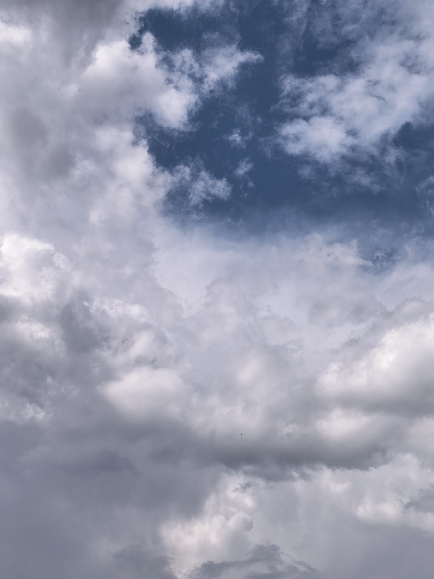 The image depicts a cloudy sky with a mix of white, gray, and dark blue clouds. The clouds are textured and appear to be moving, with some areas showing more density and shadow than others. The sky is the sole subject of the image, with no discernable man-made objects or geographical features visible. The lighting suggests it might be daytime, but the heavy cloud cover creates a subdued atmosphere.