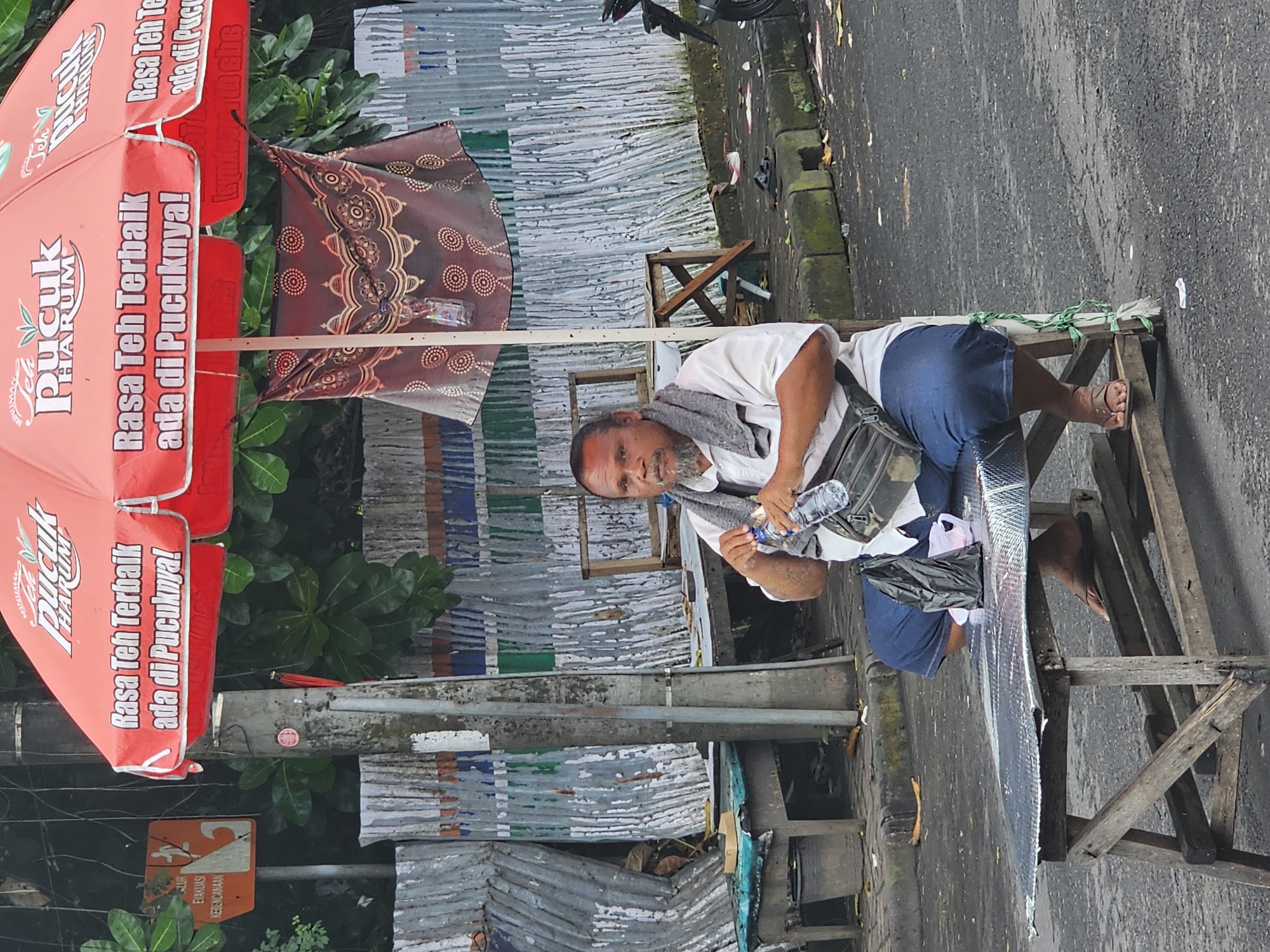 The image depicts a man sitting on a makeshift wooden structure adjacent to a street in what is likely Denpasar, Indonesia. The man is wearing a white shirt and blue shorts, with a gray scarf around his neck and a satchel bag over his shoulder. He is holding a water bottle in his hands. Beside him is a shiny surface and a black plastic bag. A red Pucuk Harum umbrella stands in the background, and nearby there is signage related to emergency evacuation. The background consists of corrugated metal sheets and various other objects, creating a cluttered urban environment.