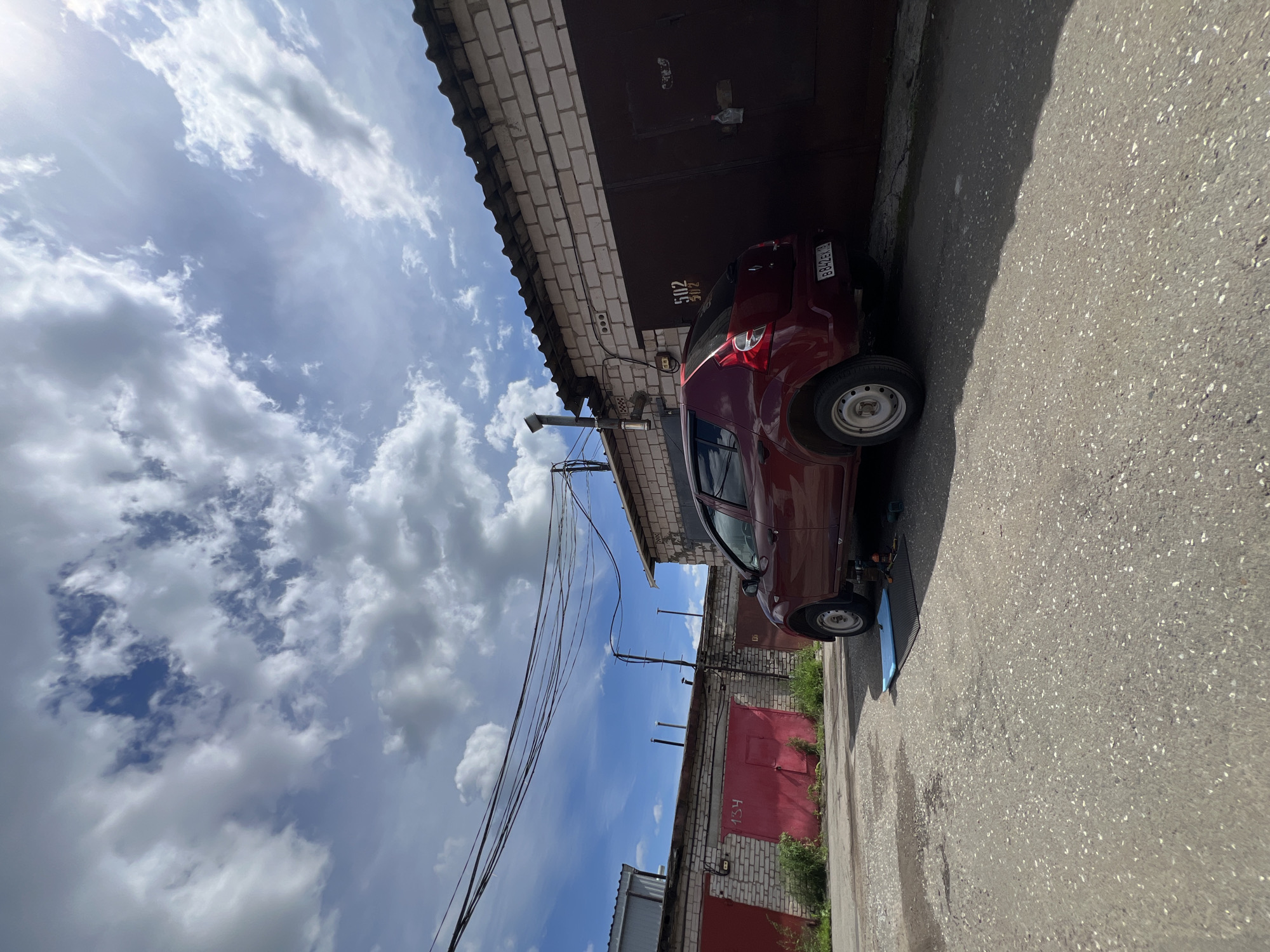 A red four-door sedan is parked on an asphalt surface in front of a row of garages. The car is positioned at an angle, facing towards the right side of the frame. Underneath the rear passenger side of the car, a blue mat and a black textured mat are laid out on the ground, with tools scattered on them, suggesting some form of repair or maintenance is being performed on the vehicle. The garages are constructed from light-colored bricks and have large, dark-colored doors. Numbers are visible on some of the garage doors, including "502" and "502" on one and "134" on another red garage door to the left. Overhead, a network of electrical wires is strung across the sky. The sky is blue with scattered white clouds. The environment appears to be an outdoor parking area or a service area within a garage complex, likely in the city of Veteran-2, Russia.